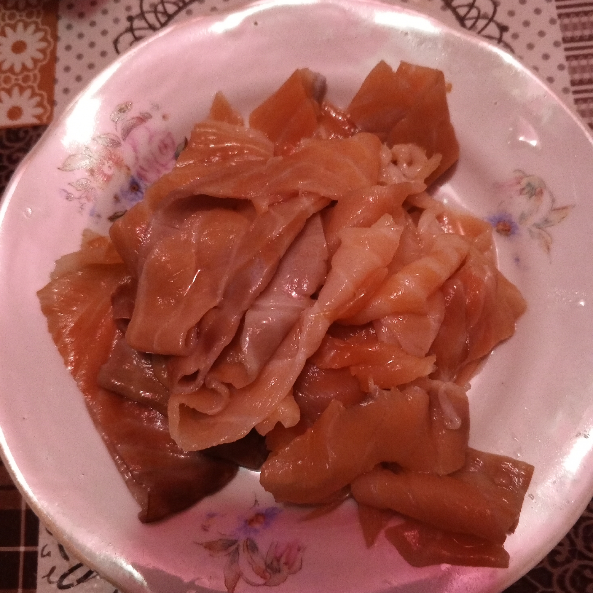 A close-up, overhead shot shows a white, floral-patterned plate filled with thinly sliced, raw salmon. The salmon pieces are a pale orange-pink color and have a slightly translucent appearance, with some pieces appearing more cooked and firm than others. They are piled loosely in the center of the plate, with some pieces overlapping and others more spread out. The plate has a delicate floral design with pink and purple flowers and green leaves around the rim. The background is a blurred tablecloth with a dotted and striped pattern. The lighting is warm and appears to be artificial, casting a pinkish hue over the entire image. No people are visible in the frame. The focus is entirely on the food and the plate.