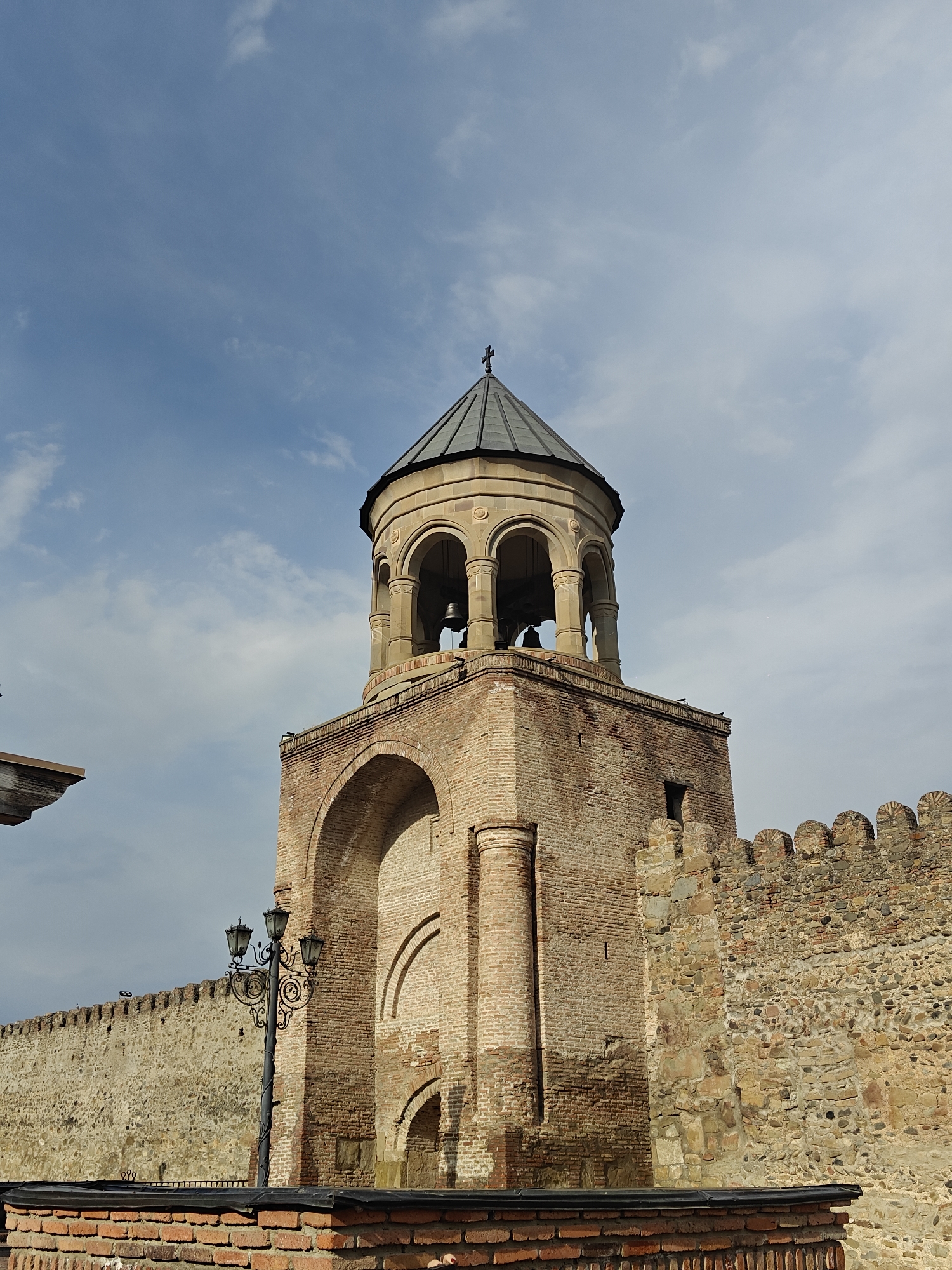 This media file captures a low-angle view of a historic bell tower and an adjoining fortified wall under a partly cloudy sky. The central subject is a robust, multi-layered structure constructed primarily from brick and stone masonry, exhibiting signs of age and restoration.
The bell tower, positioned slightly right of center, features a lower rectangular section of weathered brick. This section includes a large, recessed archway on its visible face and a thick, rounded column on its left side. Above this, a more ornate, lighter-colored stone section houses the bell chamber, characterized by a series of arched openings supported by slender columns. Within these arches, dark bells are visible. The tower is capped with a dark, conical roof, which culminates in a small cross at its apex, indicating a religious function.
To the right of the tower, a thick, crenellated wall extends horizontally across the frame. This wall is constructed from a mix of irregular stones and smaller bricks, suggesting multiple phases of construction or repair. The crenellations on the top edge of this wall are prominent. A small portion of a similar wall, made entirely of stone, is visible to the left foreground.
A decorative, black metal street lamp with multiple lanterns stands in front of the lower brick section of the tower, providing a contrast with the ancient architecture. The sky in the background is a light blue with scattered white, wispy clouds, suggesting daytime conditions, likely either morning or late afternoon given the soft lighting. No people or visible text are present in the image. The overall scene conveys a sense of historical grandeur and architectural resilience.
