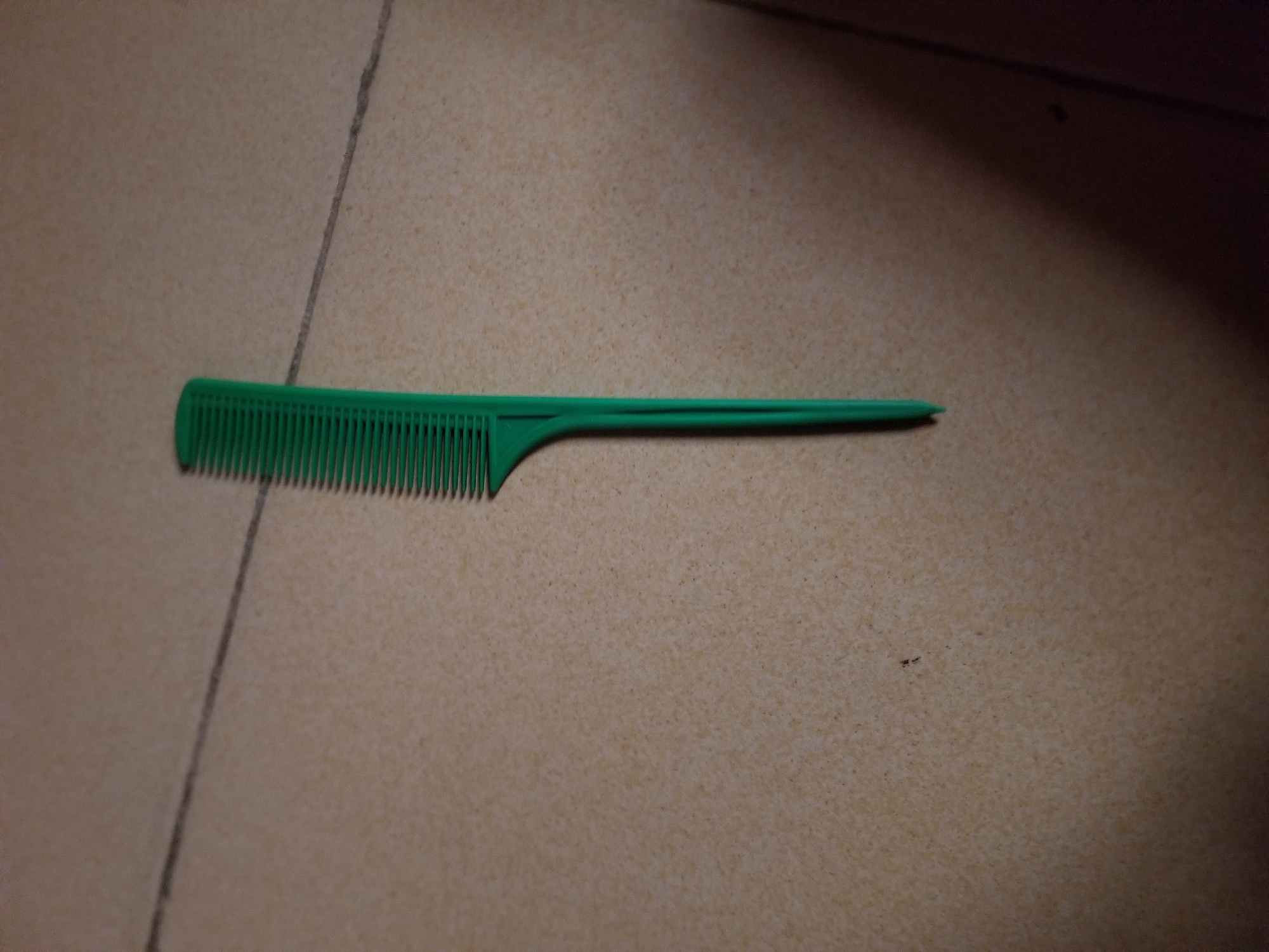 A green comb is placed on a tiled floor. The tiles are beige with dark grey grout lines. One grout line runs diagonally from the top left to the bottom right of the image, passing behind the comb. The comb is oriented vertically, with its teeth facing upwards and to the left. The handle of the comb tapers to a point. The background is mostly out of focus, with some shadow visible on the right side of the image. There is a small dark speck on the tiles in the lower left quadrant of the image. The scene appears to be indoors, likely in Jauro Yinu, Nigeria.