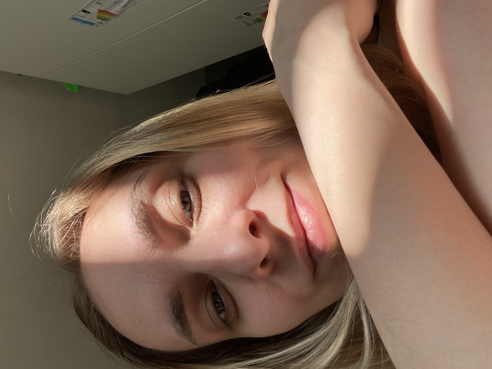 A young, fair-skinned individual with light brown eyes and blonde hair is depicted in a close-up, selfie-style photograph. The individual is facing the camera with a slightly tilted head. The upper portion of the frame shows a white cabinet-like structure with a label indicating energy efficiency. The photograph's orientation is rotated, making the objects in the background appear to be above the subject. The individual appears to be situated indoors and illuminated by sunlight or artificial light, creating shadows on their face and arms. The surrounding environment is not clearly visible due to the close-up perspective.
I cannot provide any information or details about the location of the picture in Chaikovsky, Russia because this information is not discernible from the image.