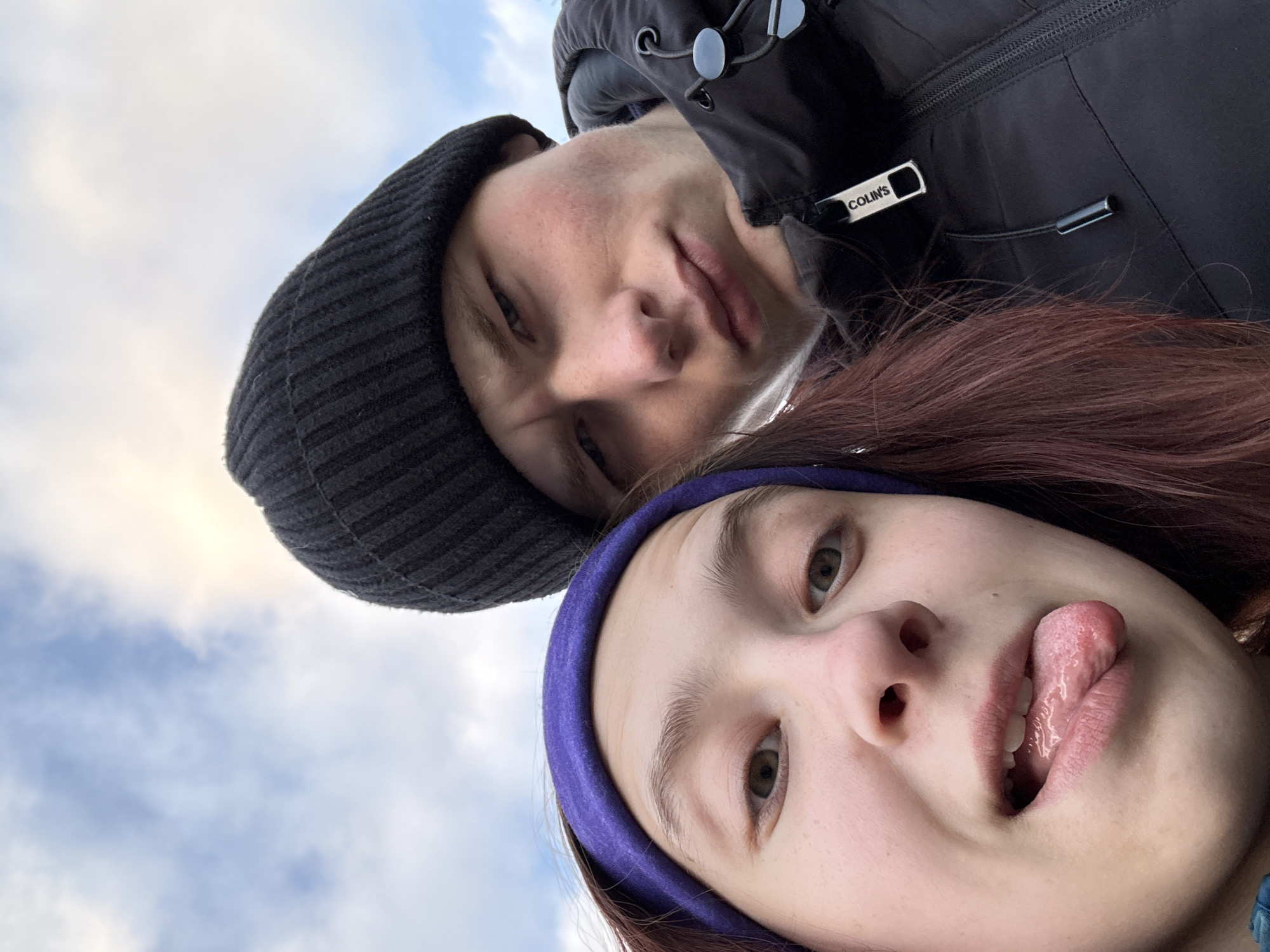 A selfie taken from a low angle features two individuals against a backdrop of a cloudy, blue sky. The person on the left, wearing a black knit cap and a black jacket with a "COLIN'S" brand tag visible on the zipper, is positioned higher in the frame. The person on the right, with reddish-brown hair and a blue headband, sticks out their tongue. The location could be Perm, Russia, but this cannot be verified from the image.