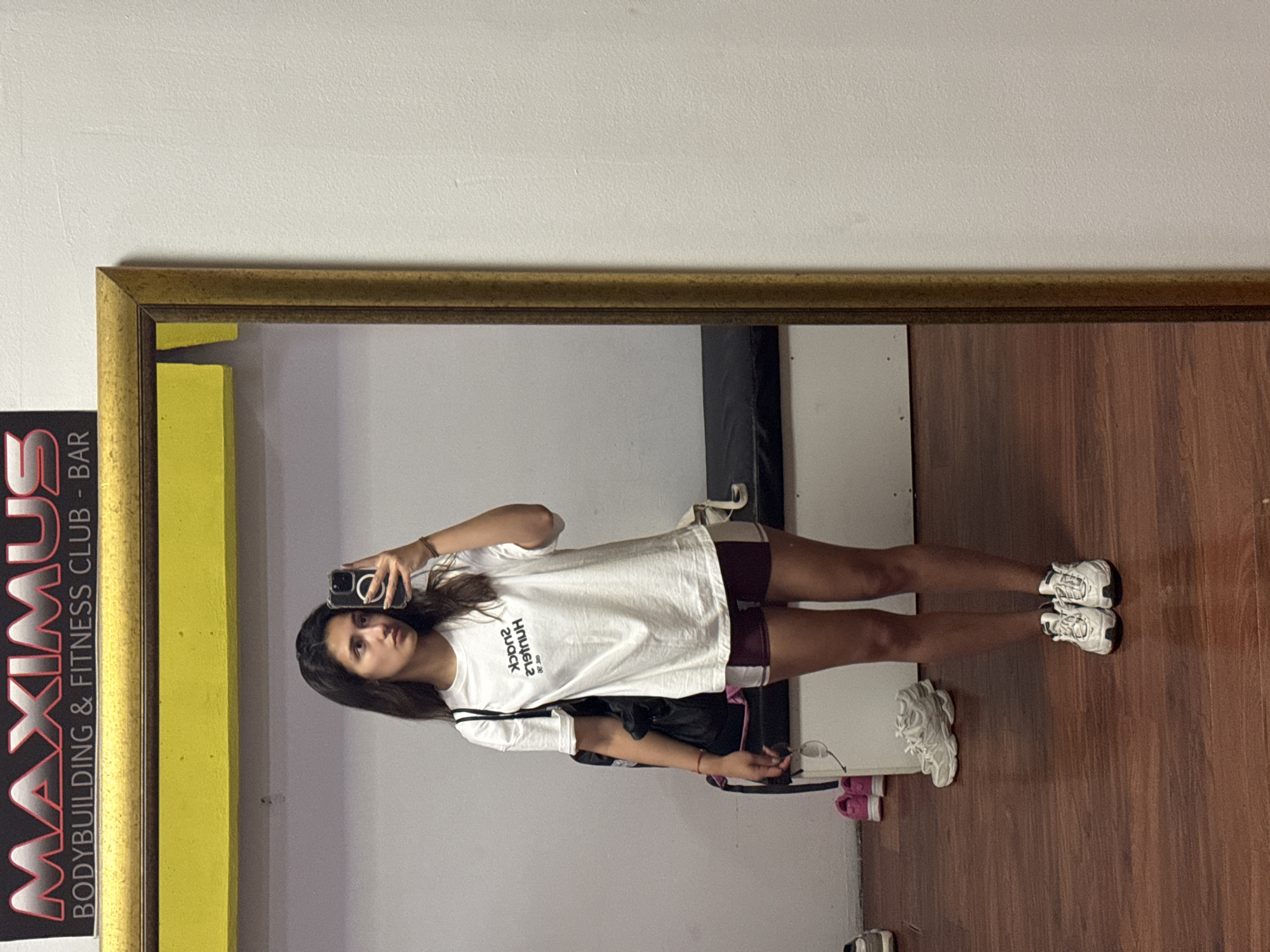 A woman is taking a mirror selfie in a gym in Bar, Montenegro. The gym is identified by a sign stating "MAXIMUS BODYBUILDING & FITNESS CLUB - BAR". The woman is wearing a white t-shirt with writing, burgundy shorts with a white stripe, and white sneakers. She is holding a cell phone in her right hand. The floor is made of brown wood. Other objects visible include other shoes and gym equipment. The mirror has a gold frame.