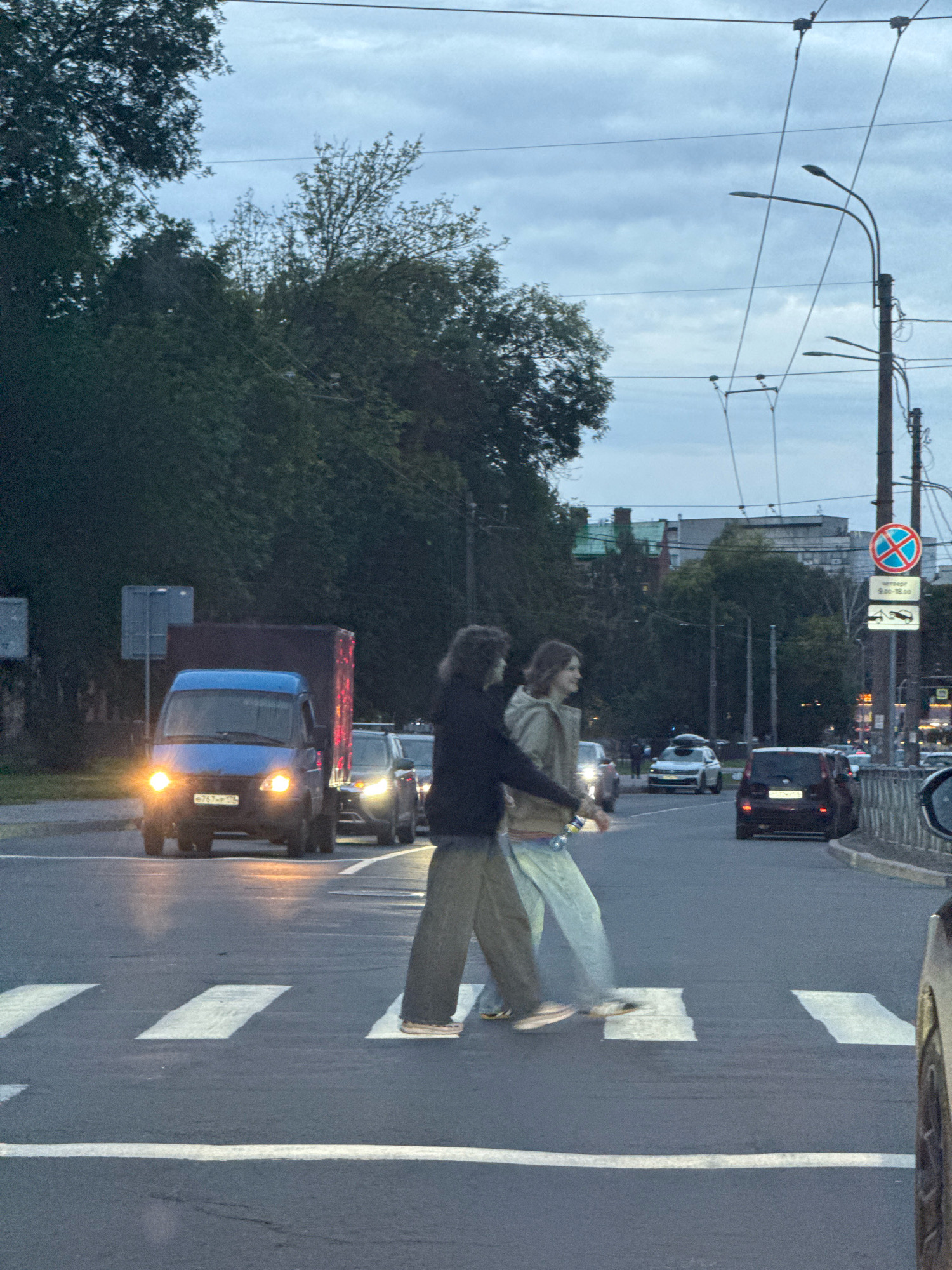 In a city street scene in an unknown location in Russia, two individuals are crossing a pedestrian walkway at dusk. The sky is overcast and blue-grey. The two people are walking side-by-side, both in casual clothing. The person on the left has long, dark hair and is wearing a dark jacket over a light-colored shirt and loose-fitting tan pants. They are holding a clear plastic bottle. The person on the right has lighter, medium-length hair and is wearing a hooded jacket with a light-colored interior and light-colored pants. They are also wearing white sneakers with stripes.
The pedestrian crossing is marked with white stripes on dark asphalt. In the background, several vehicles are visible on the road. A blue van with its headlights on is in the left lane, followed by other cars. Further down the street, a dark-colored SUV and a white sedan with a roof rack are also present. Overhead, power lines and streetlights are strung across the scene. Trees line the side of the road, and buildings with pitched roofs are visible in the distance. A traffic sign with a red circle and a blue cross inside is mounted on a pole on the right side of the road, along with other regulatory signs. The overall lighting suggests it is either early morning or late evening.