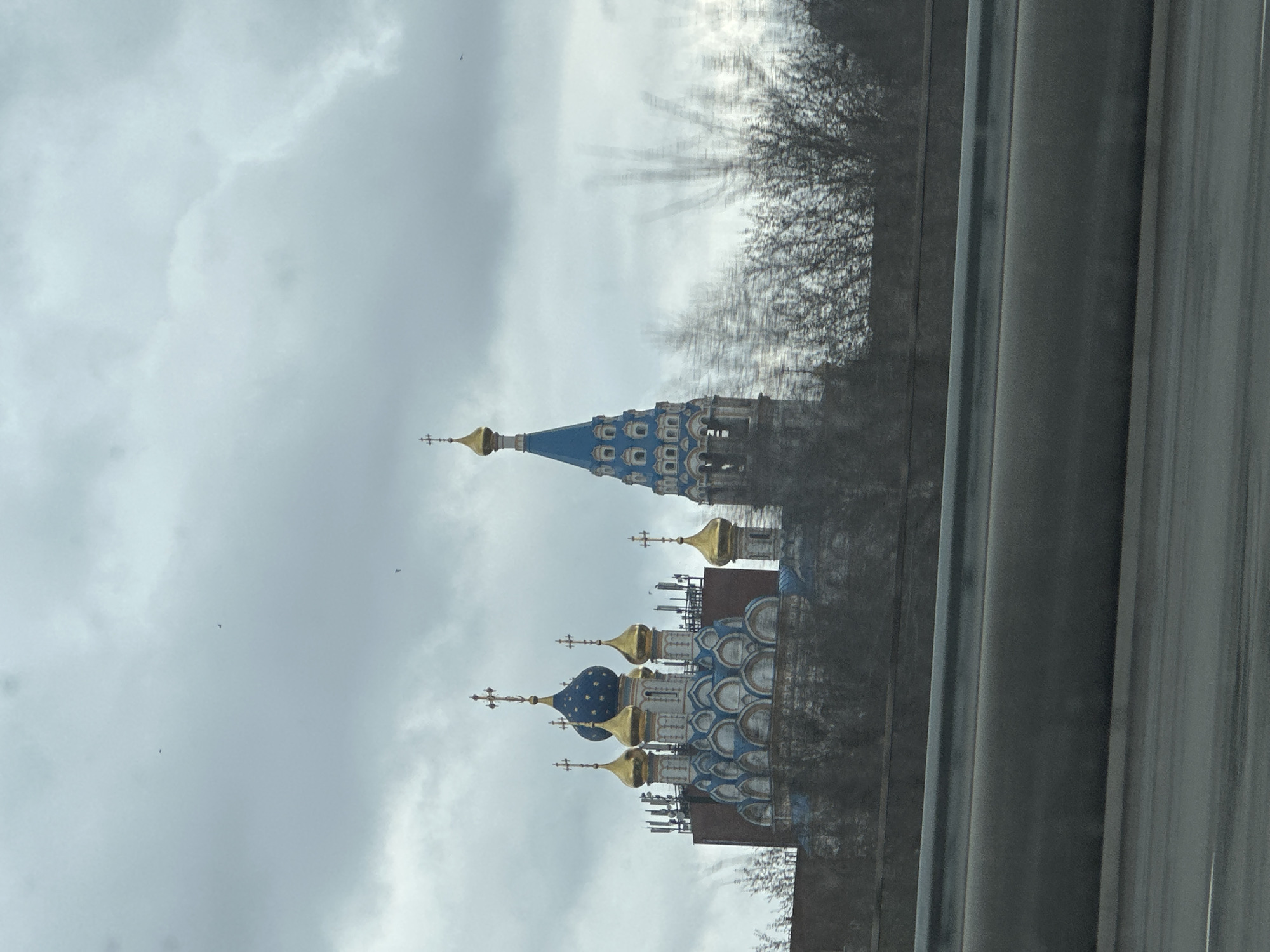 The image depicts a Russian Orthodox church viewed from behind a window, likely a car window given the vertical metallic bars visible in the foreground. The church is identifiable by its characteristic blue and gold domes topped with Orthodox crosses. The architecture suggests a Moscow location, potentially near the Kremlin, given the historic style of the building. The sky is overcast, and the environment appears urban, with bare trees visible. The angle and obscuration from the window suggest the photo was taken while in transit. There are no people distinctly visible in the image.