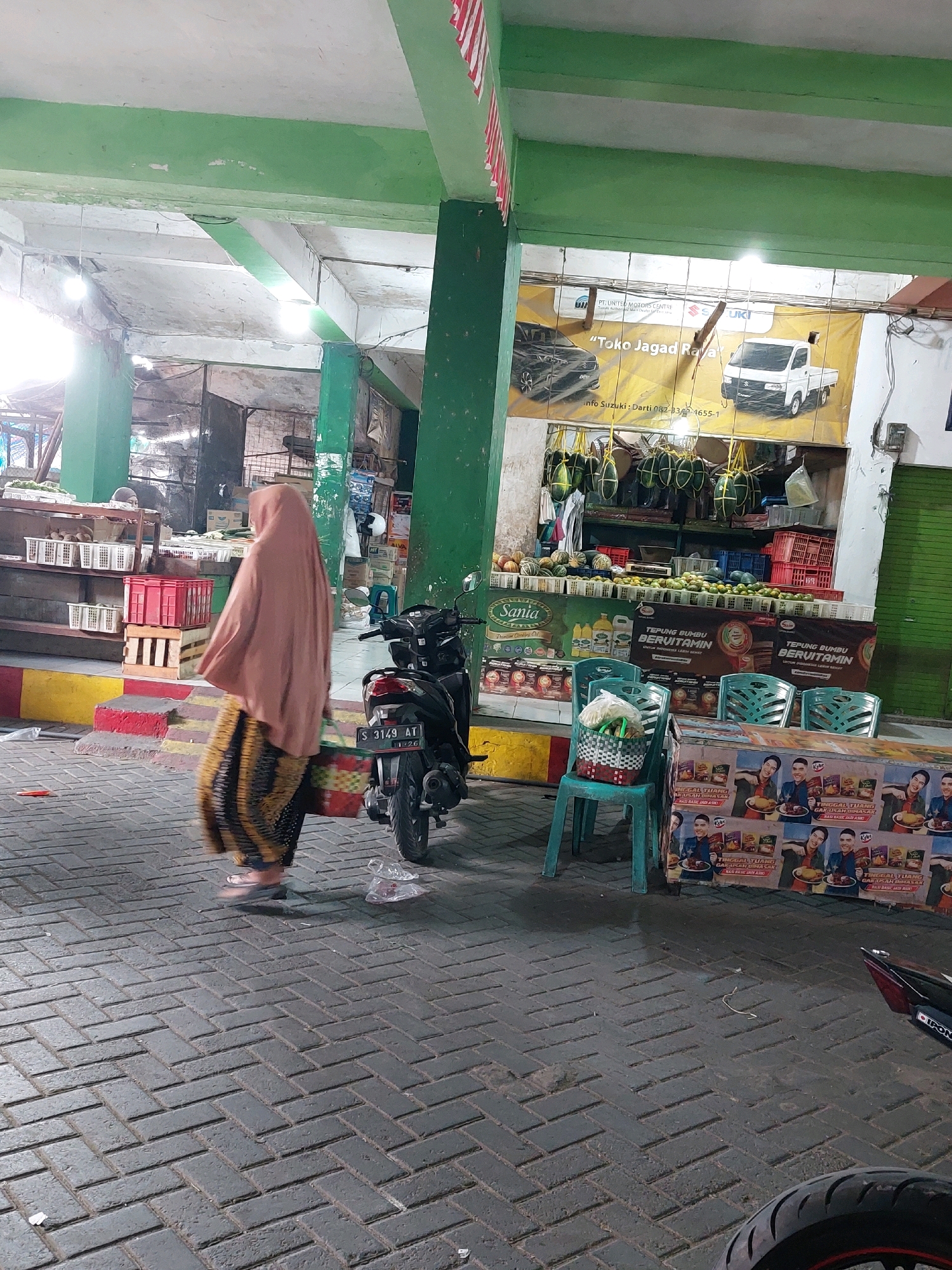 The image depicts an outdoor market scene in Sumuragung, Indonesia. A woman wearing a pink hijab and a patterned skirt walks across a brick-paved area. To her right, a black motorcycle is parked. In the background, market stalls are visible, displaying various goods including fruits, vegetables, and packaged food items. A banner advertising Suzuki vehicles hangs above one of the stalls. The market appears to be covered by a green-painted structure with exposed beams and lighting fixtures. The overall impression is of a bustling local marketplace.