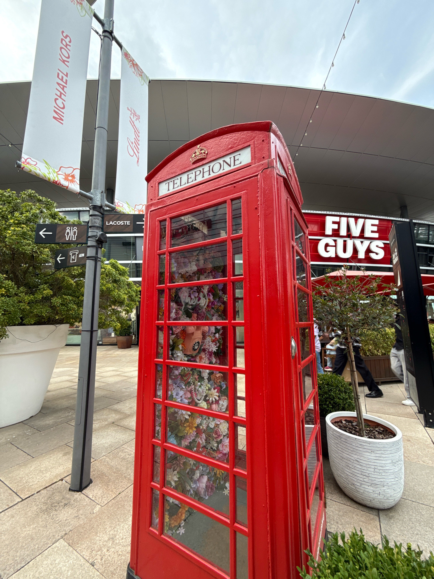 The image depicts an outdoor scene in Wolfsburg, Germany, likely within a shopping or entertainment complex. In the foreground, a classic red British telephone box is prominently displayed, its glass panels revealing a vibrant floral arrangement inside and a vintage pink telephone. To the left, a grey lamppost holds several signs, including one for "MICHAEL KORS" and another with "Lindt." Another sign indicates directions to "LACOSTE," restrooms, and an ATM. A large white planter with a lush green bush stands beside the lamppost. In the background, the red facade of a "FIVE GUYS" restaurant is visible. The ground is paved with light-colored stones. The sky is overcast. Several individuals are partially visible in the background, suggesting this is a public area.