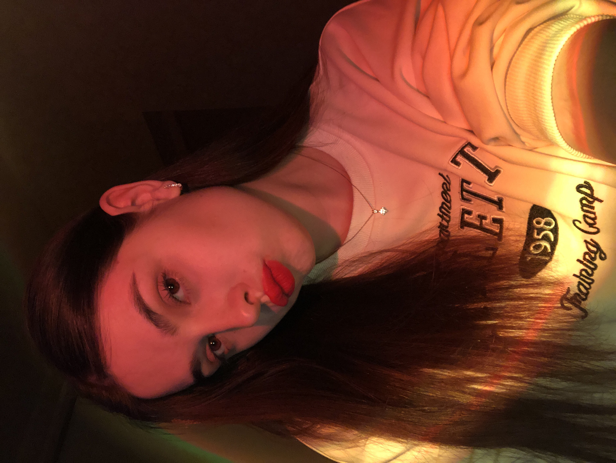 A head-and-shoulders portrait of a young woman, rotated 90 degrees clockwise. She has long, dark brown hair that cascades over her shoulder. Her face is prominently lit by a strong red light, highlighting her fair complexion, bold red lipstick, dark eyebrows, and brown eyes. A silver-toned ear cuff with small gems is visible on her upper left ear. Around her neck, she wears a delicate chain necklace featuring a small, sparkling, possibly star-shaped or cross-shaped pendant.
She is dressed in a light-colored, likely white or off-white, crewneck sweatshirt. Dark text is visible on the sweatshirt, partially reading "partmeet", "ETI", "1958", and "Training Camp". The right side of her body and hair are illuminated by a warm orange and yellow light. The background is dark and indistinct, suggesting an indoor setting. She gazes directly towards the viewer with a neutral expression, her head tilted, creating a dynamic pose. The dramatic colored lighting gives the image a stylized and moody aesthetic. The scene lacks specific location cues beyond the general context of Noviy, Russia.