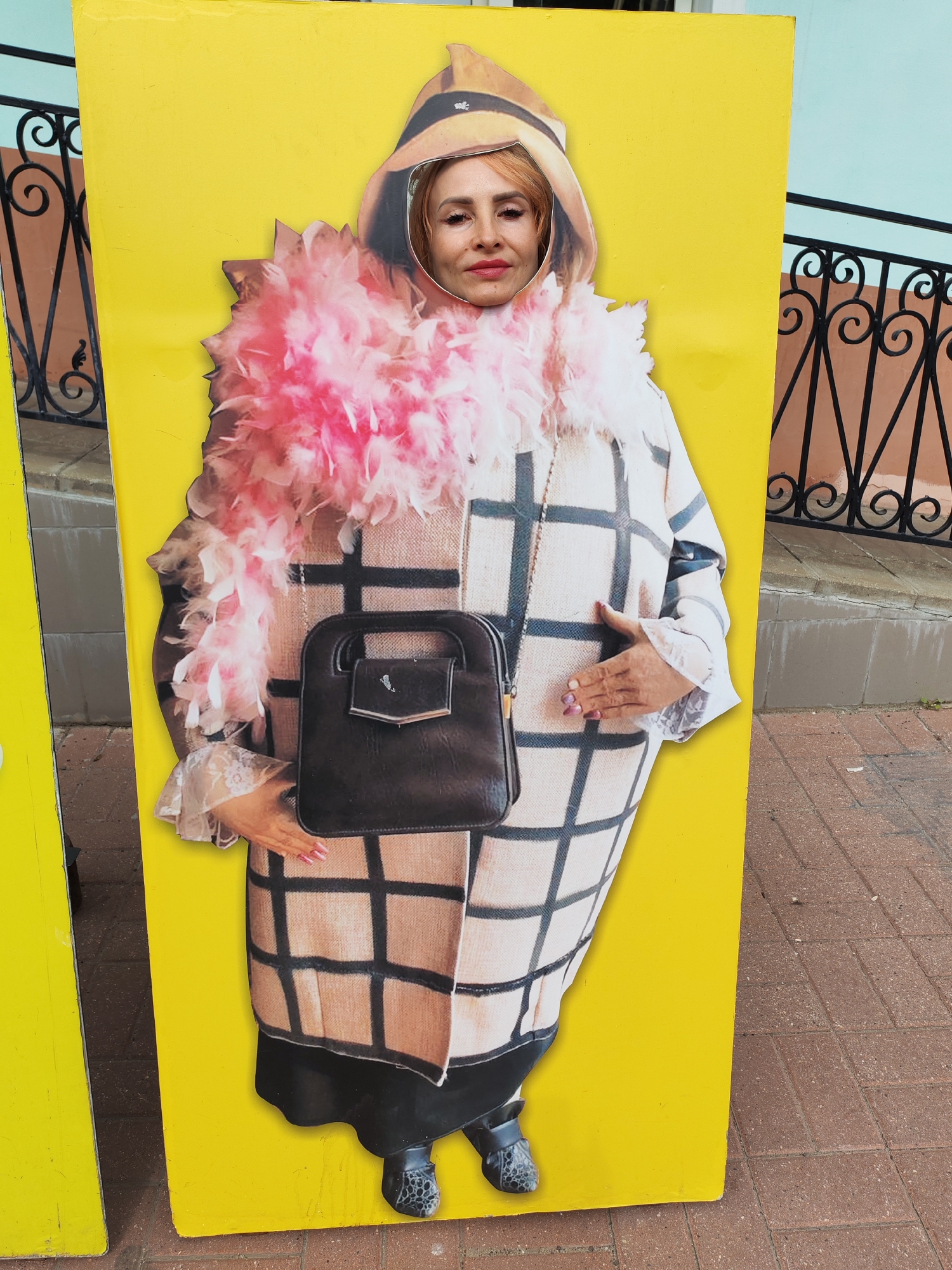 A woman's face is visible through a cutout in a yellow cardboard standee designed to resemble a full-body figure. The standee depicts a person wearing a patterned coat, a pink feather boa, and a hat. A black handbag hangs from the figure's shoulder. The standee is placed outdoors on a paved area, with a decorative black metal fence visible in the background. The environment suggests an urban or town setting.