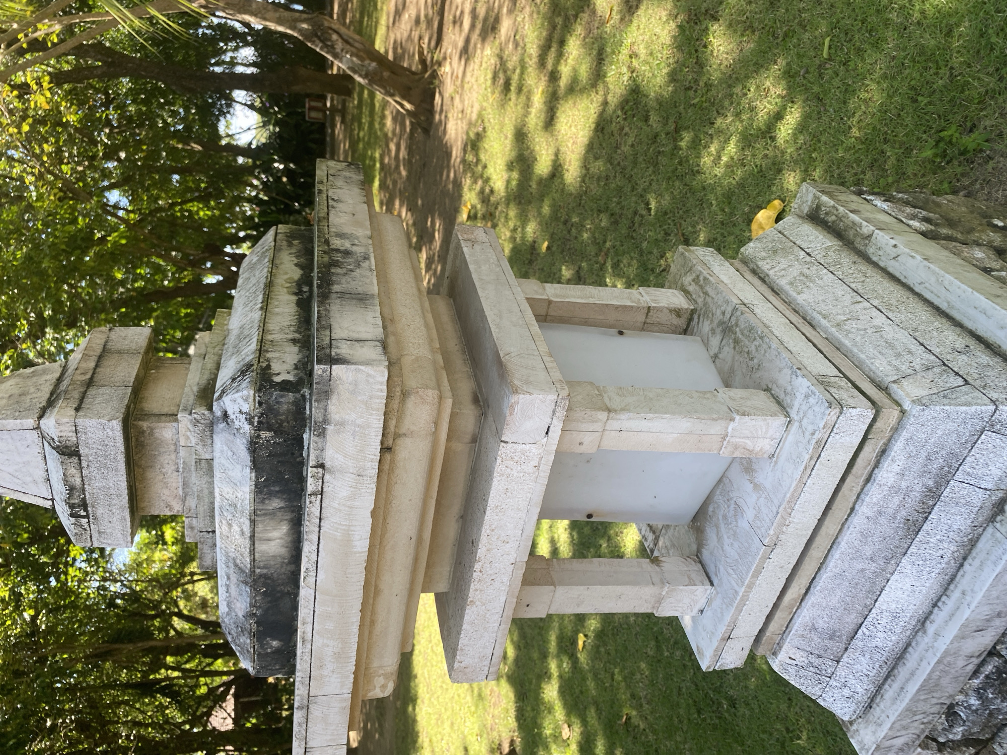 The image shows a stone pagoda structure in a grassy outdoor location, most likely in a park or garden. The structure is layered, with stacked stone elements decreasing in size towards the top, and has a central, white, box-like component. The stone has a weathered appearance, with gray and white coloration, and some areas show darker staining. Green grass and trees are visible in the background, with dappled sunlight filtering through the trees. The image is taken from a low angle, looking upwards towards the pagoda. Based on the prompt, this is in Kuta Selatan, Indonesia.