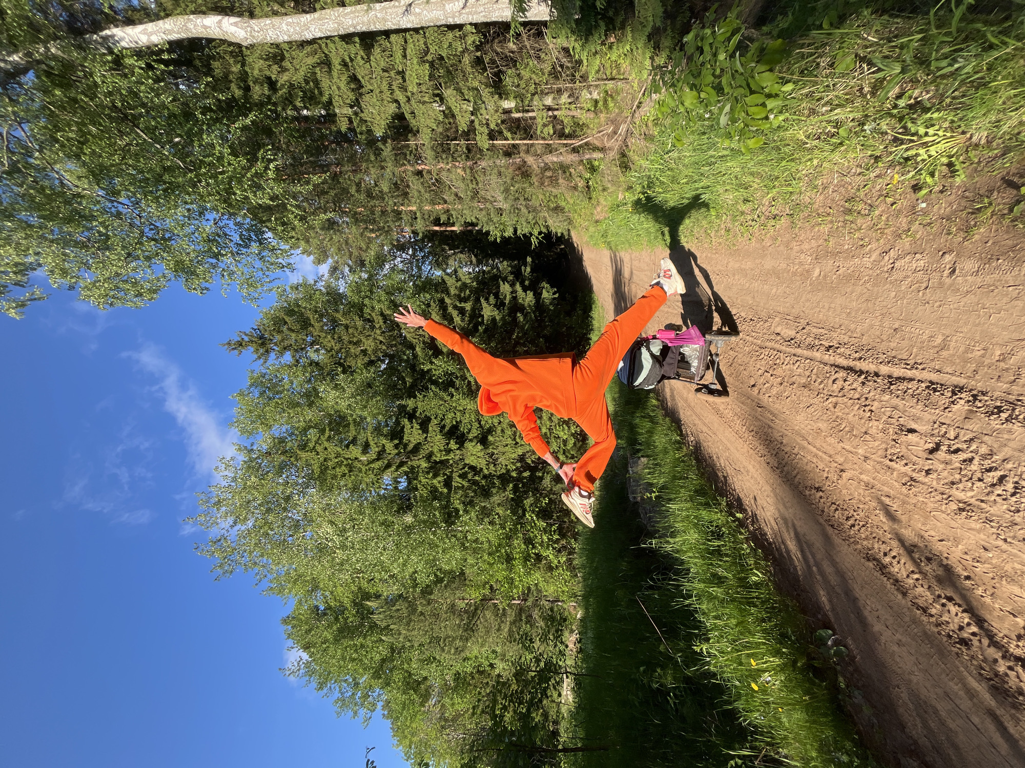 The image, intentionally rotated 90 degrees clockwise, captures a dynamic outdoor scene in what appears to be a forest or wooded park in Chaikovsky, Russia. The central subject is a person in mid-air, seemingly captured during a jump or an energetic pose. They are dressed in a vivid, solid orange hooded jumpsuit and light-colored athletic shoes, with their arms stretched wide and legs split apart, conveying a sense of freedom and motion.
The person is positioned above a clear dirt path or road that runs through the frame. The path is light brown, showing textures of soil and possibly tire tracks. On the ground next to where the person is jumping, a backpack or bag, colored blue and green with some pink elements, is visible, suggesting a temporary stop or a break from an activity.
The setting is lush and green, characteristic of a natural environment. Tall trees, including both light-barked deciduous trees like birch and darker coniferous trees, flank the path, creating a dense backdrop of foliage. Green grass and smaller plants grow along the edges of the path. The sky above is a clear, bright blue with a few scattered white, wispy clouds, indicating fair weather. Strong sunlight illuminates the scene, casting distinct shadows on the ground, suggesting it is daytime, possibly late morning or afternoon. No visible text is present in the image.