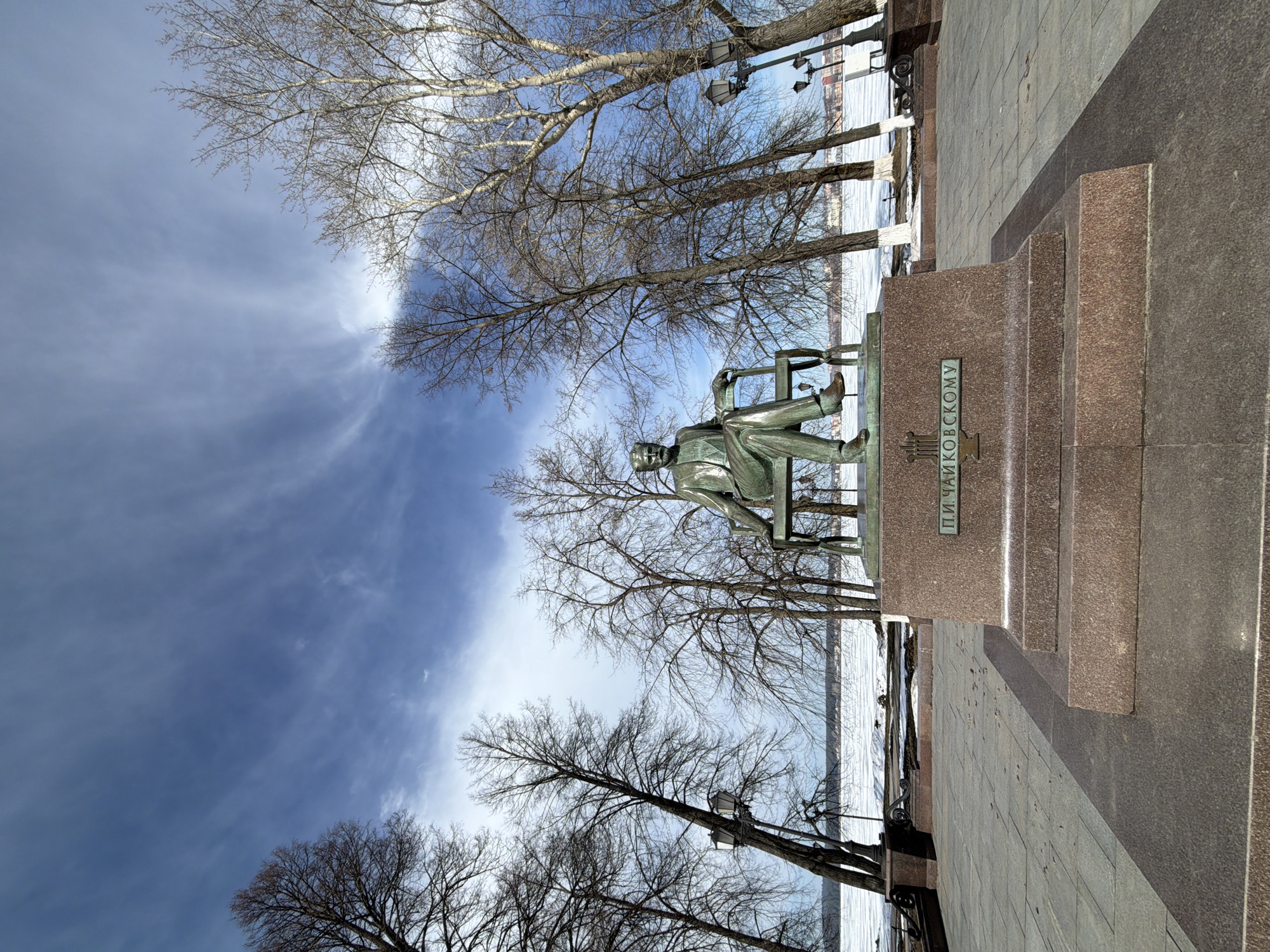 The image shows a statue of Pyotr Ilyich Tchaikovsky in Votkinsk, Russia. The statue depicts Tchaikovsky sitting on a chair. The base of the statue has the inscription "P.I. Tchaikovsky." The statue is positioned near a body of water, which appears to be partially frozen. Bare trees surround the statue, and street lamps are visible along the waterfront. The sky above is blue with wispy clouds. The surrounding pavement and statue base are made of stone or concrete.