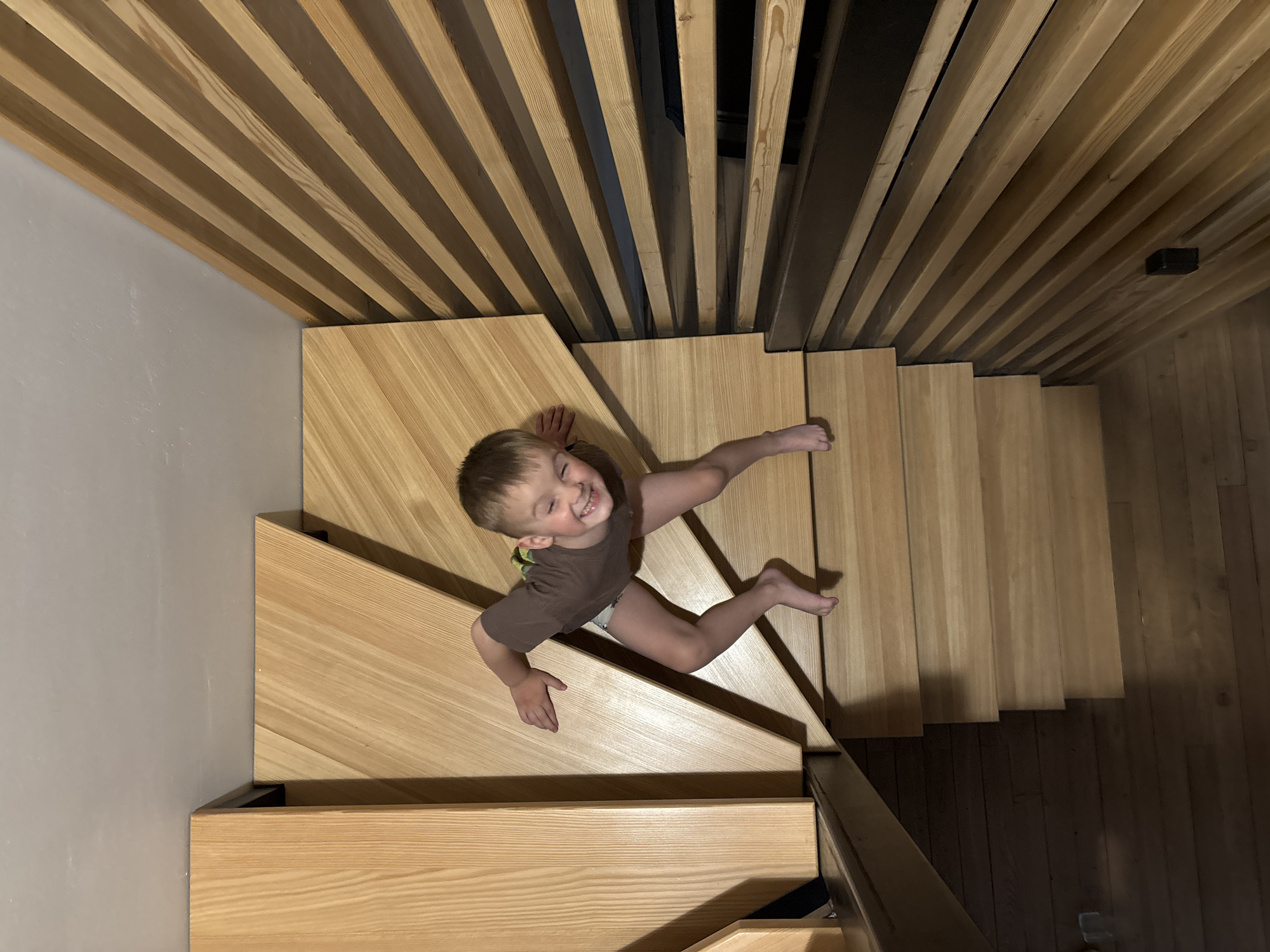 A high-angle shot shows a smiling child on a wooden staircase. The child, fair-skinned with short blonde hair, is wearing a brown t-shirt and a diaper. The child is on the lower portion of a light wooden staircase. The staircase ascends diagonally from the lower left to the upper right of the frame. Behind the staircase are vertical wooden slats. The background includes a light gray wall to the left and a dark wooden floor to the right. There is no verifiable evidence of location in the city of Chaikovsky, Russia.