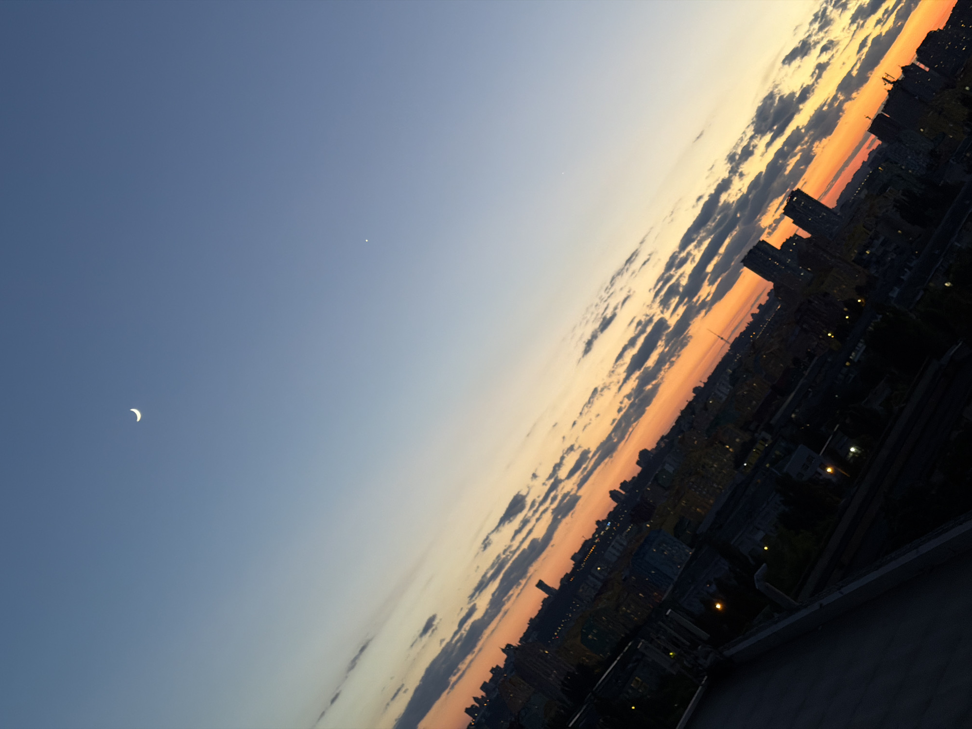 The image depicts a tilted view of Kyiv, Ukraine, during a sunset. The sky is a gradient of deep blue at the top, transitioning through lighter blues, yellows, and oranges near the horizon, with scattered clouds. A crescent moon is visible in the upper left quadrant of the sky, and a single bright star is present in the upper right. The cityscape below is silhouetted against the vibrant sky, with buildings and structures casting long shadows. Lights are visible twinkling from within some of the buildings, indicating that evening has begun. In the foreground, a dark rooftop with some visible architectural elements occupies the lower left portion of the frame. The overall impression is of a serene urban landscape at twilight.