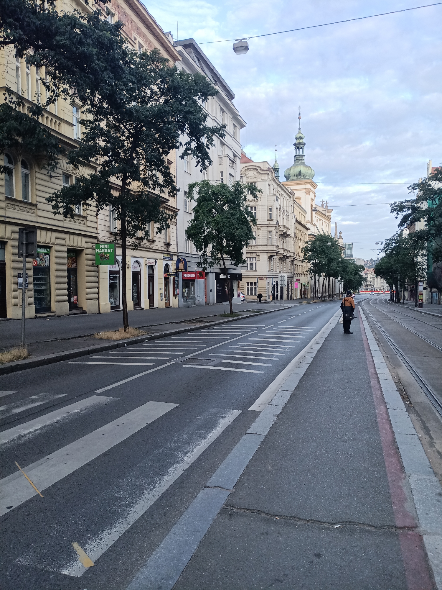 The image depicts a wide street in Prague, Czechia, likely in the city center, characterized by its historic European architecture. On the left side of the street, a row of buildings with ornate facades stands, featuring multiple stories with windows and balconies. Trees with dark green foliage line the sidewalk in front of these buildings. Shops with signs such as "MINI MARKET" and "MEGA-KNÍHY" are visible on the ground floor. The street itself is paved with asphalt, and a crosswalk with white stripes is prominent in the foreground. Tram tracks run along the right side of the street, bordered by a curb. In the distance, more buildings with distinctive spires are visible, suggesting a historic urban setting. A single person, wearing an orange jacket and dark pants, is standing on the sidewalk near the tram tracks, facing away from the camera. The sky is overcast with a light blue-grey hue, and power lines stretch across the upper portion of the frame. The overall scene suggests an early morning or late afternoon in a quiet part of the city, with minimal activity.
