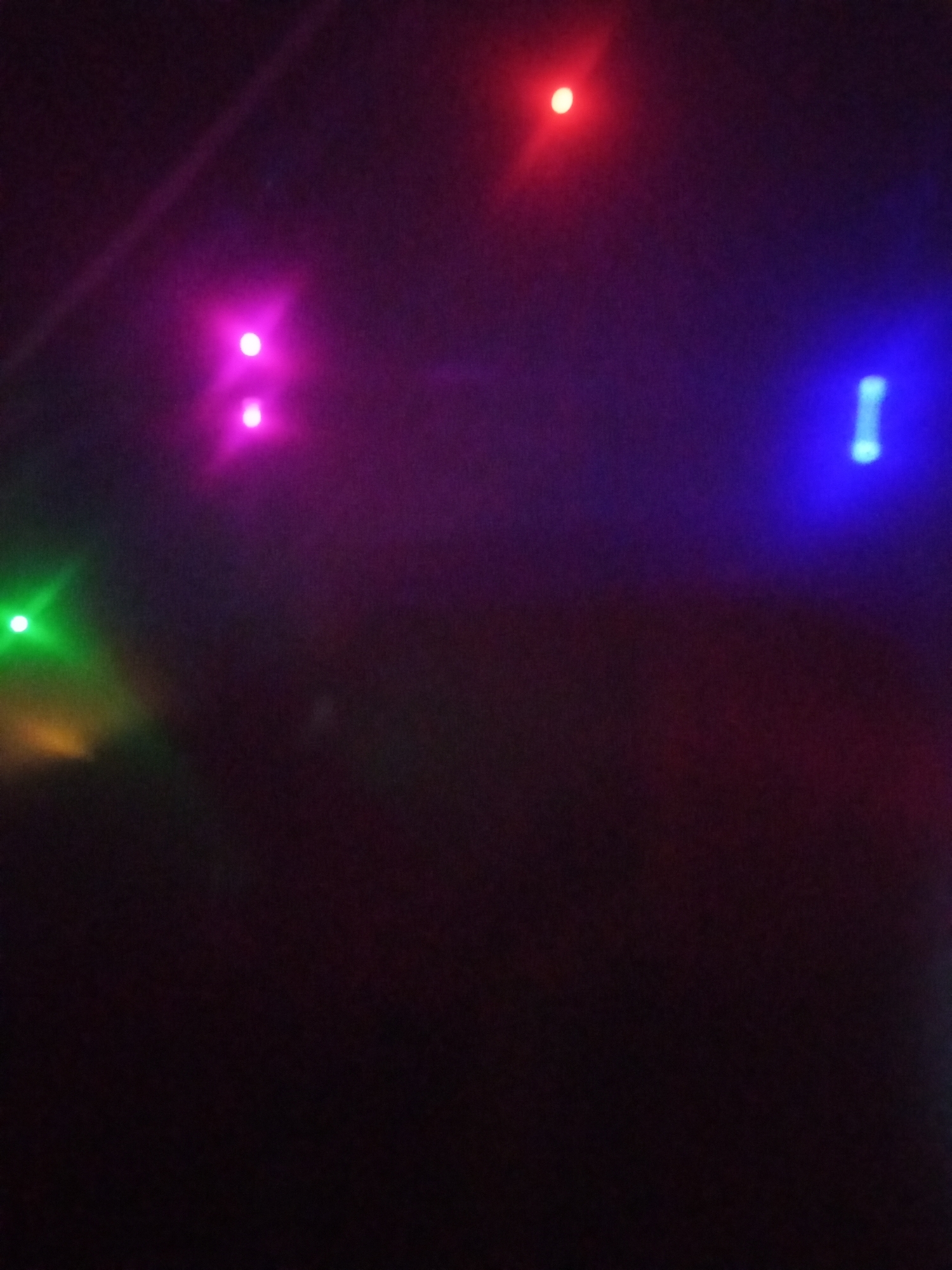 The image is a dark, out-of-focus photograph featuring several blurred, colorful lights against a black background. The lights appear to be from a string of decorative lights or possibly stage lighting. There are discernible spots of color including bright pink or magenta, red, green, yellow, and blue. The overall impression is of a dimly lit environment with prominent light sources. There are no discernible people, objects, actions, interactions, or specific environmental features that would allow for localization to Lhokseumawe, Indonesia. The image lacks sufficient detail for a forensic description beyond the presence and colors of the lights.
