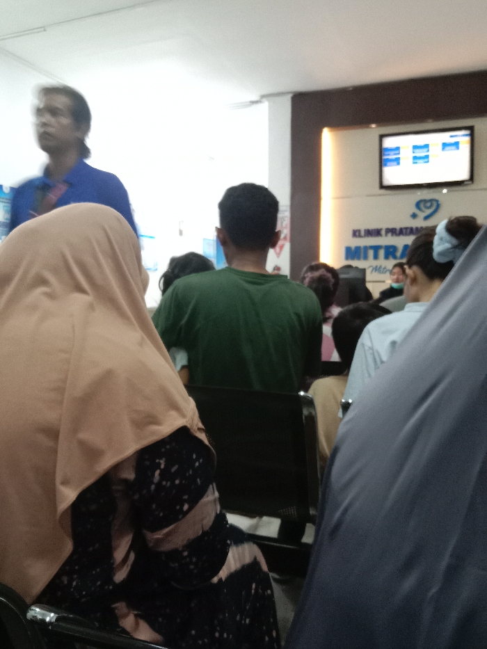 The image displays an indoor waiting area of Klinik Pratama Mitra (Primary Clinic Mitra) located in Gladaksari, Indonesia.
In the immediate foreground, a person is seated with their back largely to the camera, wearing a light brown hijab and a dark-patterned long-sleeved dress. To their right, a dark grey, draped object, possibly an umbrella, partially obscures the view of another seated individual.
In the mid-ground, a man with dark, shoulder-length hair, wearing a blue polo shirt, is captured in motion, his face blurred, standing and looking towards the left of the frame. Behind the foreground individual and slightly to the right of the man in blue, another person is seated facing away from the camera, wearing a dark green t-shirt. Further right, a seated individual with light blue hair accessories is visible from the back, wearing a light blue top. Multiple other indistinct individuals are visible throughout the mid-ground and background, predominantly seated on dark-colored chairs.
The background features a white wall with a prominent brown accent panel on the right. A flat-screen television, displaying a digital interface with blue elements, is mounted on the wall. Below the television, a sign is visible, reading "KLINIK PRATAMA" and "MITRA" with a heart logo. A female clinic staff member, wearing a medical mask, is partially visible behind a counter or desk in the background below the sign. The room is brightly illuminated by overhead artificial lighting.