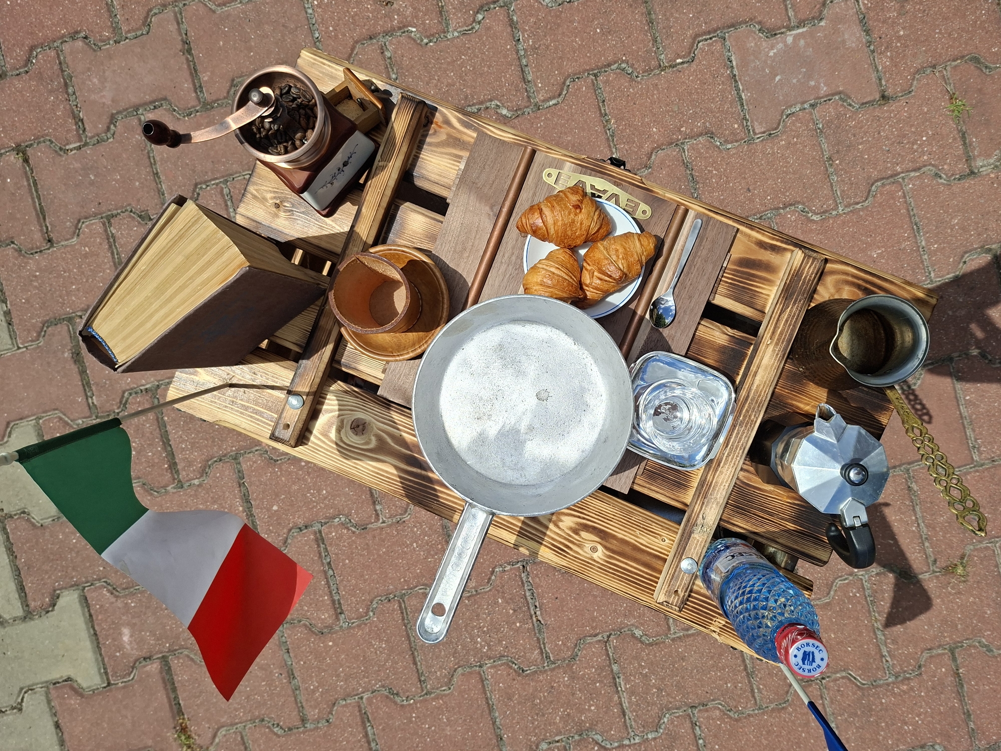 In Șerpeni, Moldova, a wooden pallet table is set up outdoors on a brick-paved surface. On the table, an Italian flag is positioned to the left, and a book is placed to the right, next to a vintage manual coffee grinder filled with coffee beans. A small wooden cup sits on a saucer. In the center of the table, a metal pan contains a frothy liquid. To the right of the pan, a plate holds three croissants, with a spoon nearby. Further down the table, a blue plastic water bottle rests next to a Moka pot. In the lower right corner, a small metal pitcher is visible. The scene is illuminated by natural daylight, casting shadows on the brick ground.