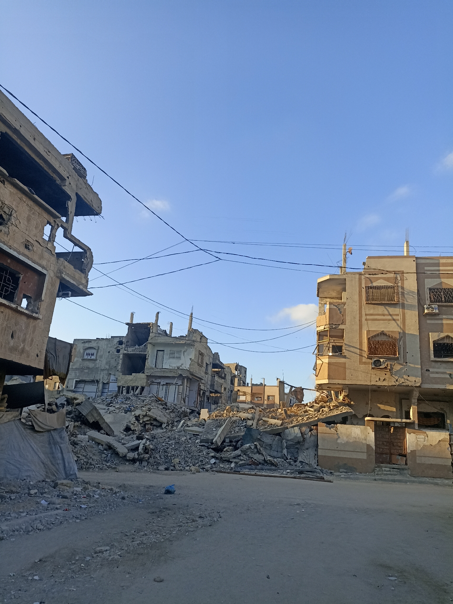 A street view in an urban area shows buildings heavily damaged by conflict. The sky is clear and blue with a few wispy clouds. In the foreground, a dirt road is covered with debris, including rocks and broken concrete. On the left, a partially destroyed building stands, its facade cracked and windows broken. A tarp is draped over some of the rubble. To the right and center, more severely damaged buildings line the street. These structures are reduced to rubble, with exposed rebar and shattered walls. Power lines crisscross above the scene. Some buildings show signs of repeated damage and repair. The overall impression is one of destruction and neglect in a war-torn urban environment.