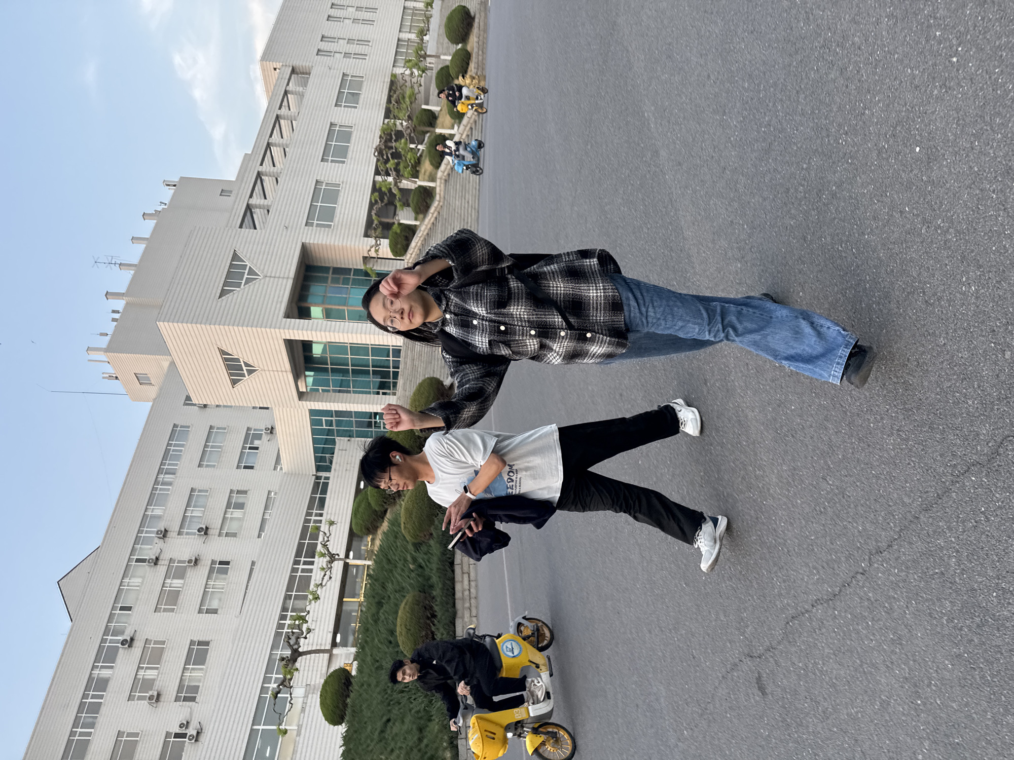 The scene shows two people posing for a picture in front of a large building in Zhongtun, China. The building has a modern architectural style with multiple windows and a white facade. The person closest to the camera is wearing a checkered shirt and jeans, while the other is wearing a white t-shirt and black pants. In the background, there are people riding yellow tricycles. The ground is paved with asphalt. The weather appears to be clear and sunny.