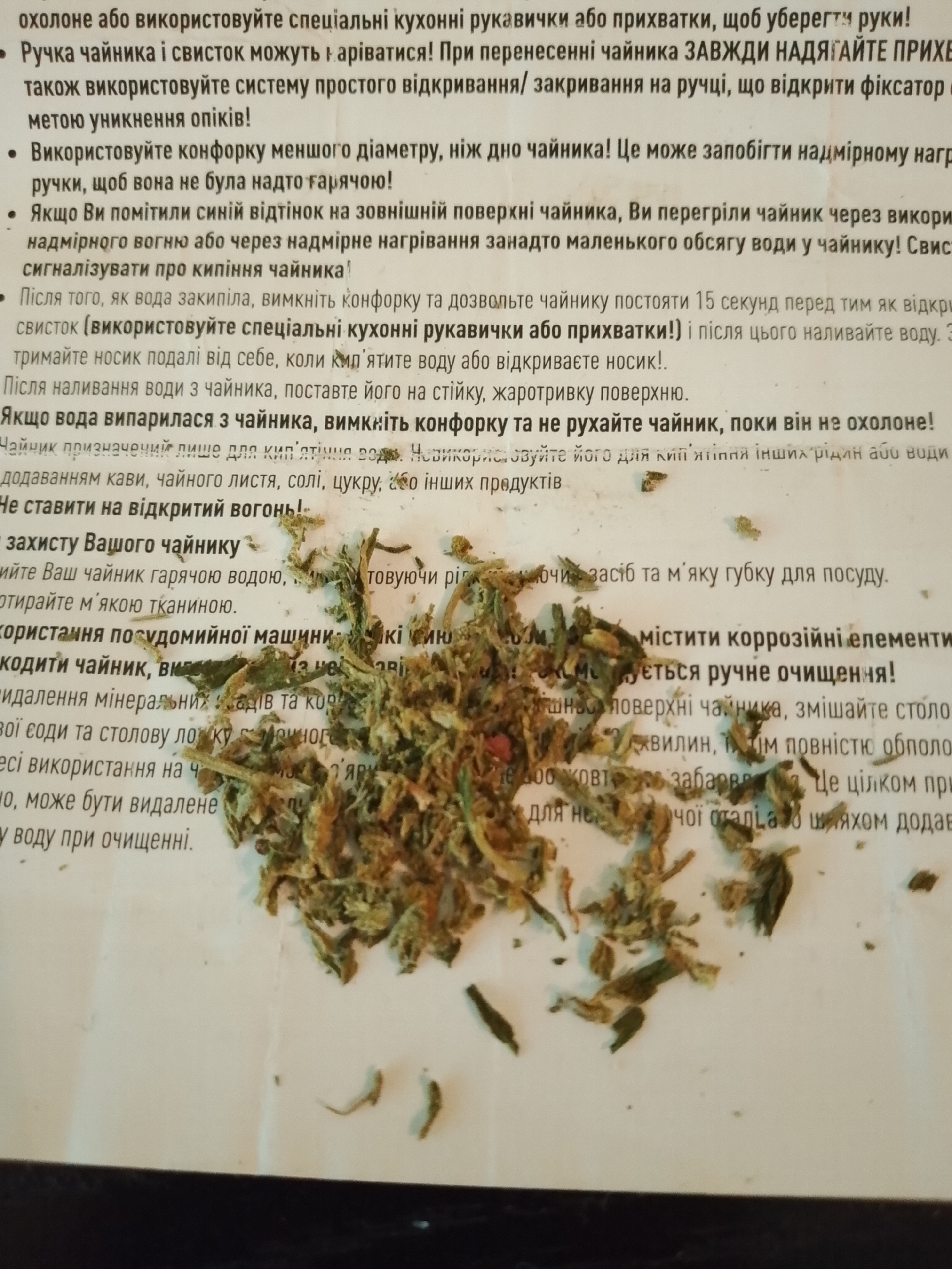 This media file is a close-up, overhead shot of a white surface with a scattering of dried herbs and a portion of text in Ukrainian. The herbs appear to be loose-leaf tea or a similar herbal blend, with various shades of green and brown. Some pieces are finely chopped, while others are larger leaves and stems. The text is printed in a black, sans-serif font and discusses the use and care of a kettle. Key points from the text include warnings about hot surfaces, proper use of the kettle's handle and whistle, avoiding excessive heat, and instructions for cleaning. There is no indication of people or a specific setting other than the white surface which might be a table. The lighting suggests it could be daytime, but there are no distinct shadows to confirm a time of day. There are no weather cues or specific location indicators within the image itself.