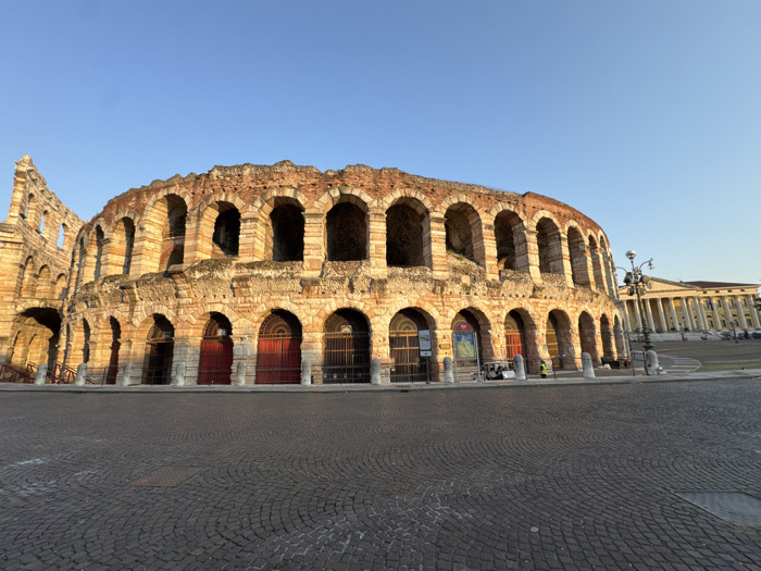 An exterior long shot depicts the Arena di Verona, a Roman amphitheater, situated in Piazza Bra, Verona, Italy, under a clear, light blue sky. The massive stone structure, constructed from reddish-brown and beige masonry, features multiple tiers of arched openings along its visible facade. Warm, low-angle sunlight illuminates the stone, casting long shadows.
The ground-level arches are fitted with various gates and doors; some are solid reddish-brown panels, others appear to be dark metal grates. Promotional posters are visible within several arched recesses on the lower level. To the far left, a section of the Arena displays more extensive ruin, with exposed internal structures.
The foreground is dominated by a large, semicircular cobblestone plaza, presenting a pattern of concentric arcs. White stone bollards line the edge of the plaza immediately in front of the Arena. A single, ornate black lamppost stands to the right of the Arena.
In the mid-ground, near the base of the Arena, a small white utility vehicle or golf cart is parked. A solitary individual, wearing a high-visibility vest, stands near the vehicle. In the background, to the right of the Arena, a large, light-colored classical building with a prominent colonnade and portico is visible across a paved area. The overall scene indicates an iconic historic urban landmark with minimal human activity.