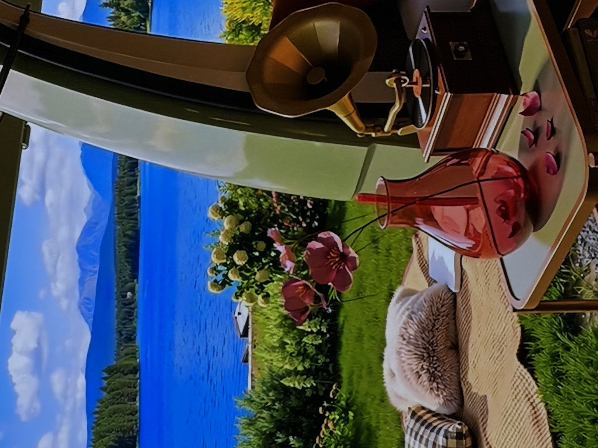 This media file presents a vibrant and serene scene, appearing to be a digital display, possibly a large television screen, exhibiting a picturesque landscape view from what seems like an interior setting. The image is rotated, but when viewed upright, it shows foreground elements on a surface, overlooking an expansive natural vista.
In the foreground, an ornate, vintage-style gramophone with a golden horn is prominent on a light-colored table or shelf. Next to it, a translucent red or reddish-pink glass vase holds what appear to be red flowers or decorative elements, with some scattered red flower petals on the surface nearby. A comfortable, fluffy light-beige pillow and a black-and-white checkered pillow are visible, resting on a textured, light brown blanket or throw, suggesting a cozy seating area within the interior space.
The background, depicted on the screen, reveals a breathtaking outdoor landscape. A vast, calm blue lake stretches across the middle ground, bordered by dense green forests and vegetation. Majestic mountains rise in the far distance, partially obscured by soft white clouds under a clear blue sky. A small, white structure is faintly discernible near the lake's edge. The foreground of the depicted outdoor scene features lush green grass and bushes with clusters of white and pink flowers.
There are no people visible in the image, nor any explicit activity taking place; it is a static, tranquil scene. The time of day appears to be bright daytime, given the clear sky and ample light. The overall mood conveyed is one of peacefulness and natural beauty, combining the comfort of an interior space with a grand outdoor view. No visible text is present in the image. The location context provided is Sofiivska Borshchahivka, Ukraine, indicating where this photograph of the display was taken.