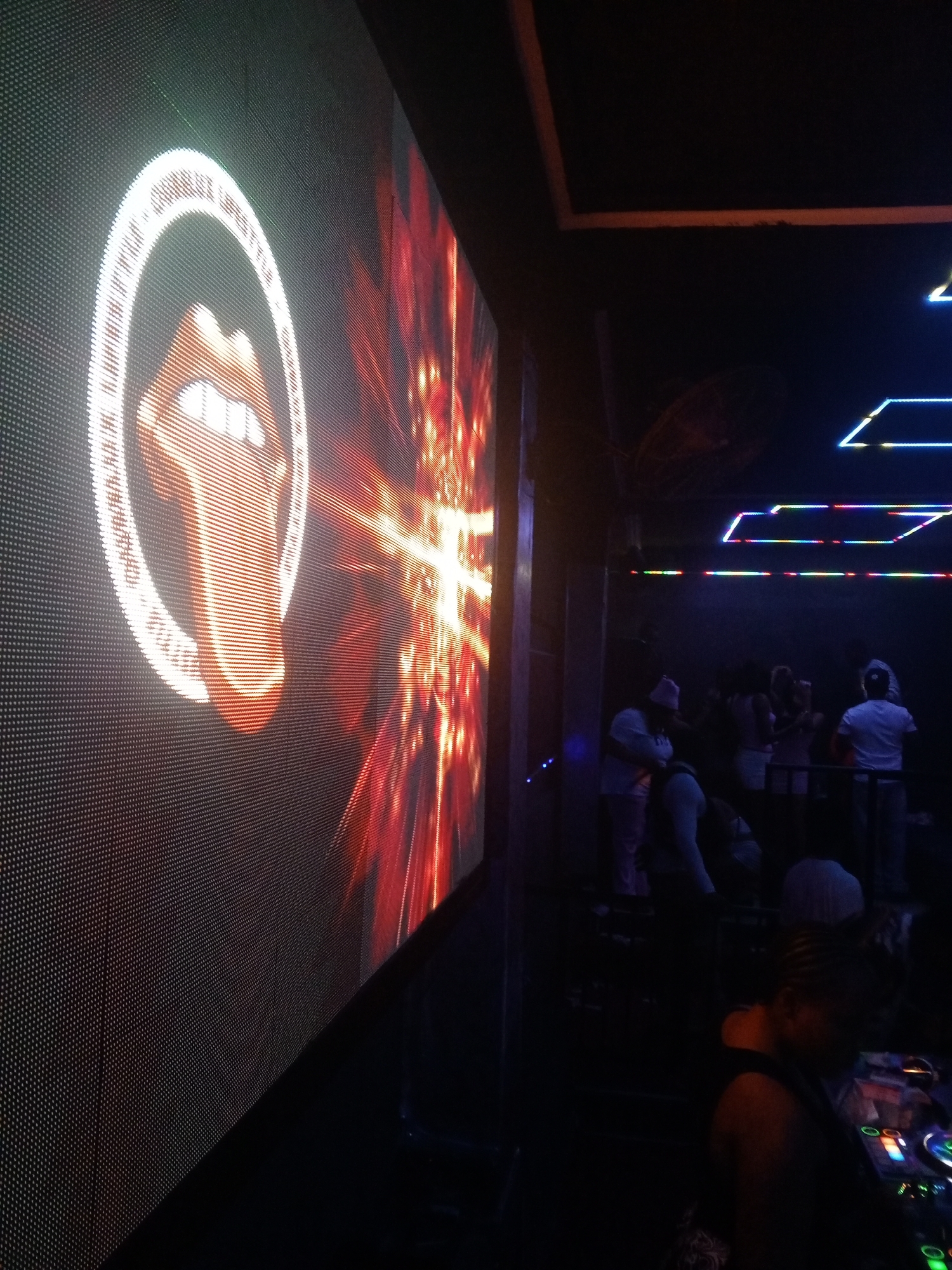 A scene in a dimly lit club in an unknown city. On the left, a large LED screen displays the Rolling Stones' logo with the words "Complex Lifestyle" around it. To the right, a partially visible crowd of people stand in the background. In the foreground, a person stands over a DJ setup with various controls and buttons visible. Above the crowd, neon lights form geometric shapes on the ceiling. The environment suggests a lively, nighttime atmosphere.