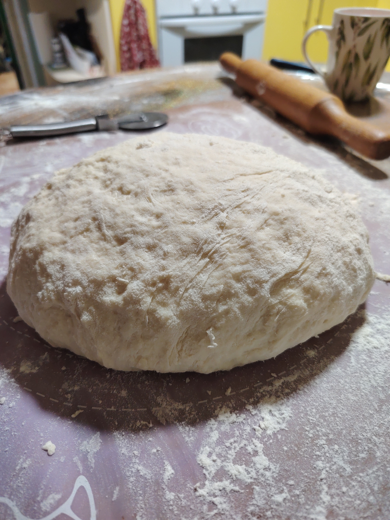 A ball of lightly floured dough rests on a floured surface with a purple grid pattern. To the left of the dough is a pizza cutter, and further to the left, a rolling pin. In the background, a mug with a leaf pattern sits on a tray. A kitchen counter and a yellow wall are visible behind the objects. The scene is indoors, likely a kitchen, in Rostov-on-Don, Russia.
