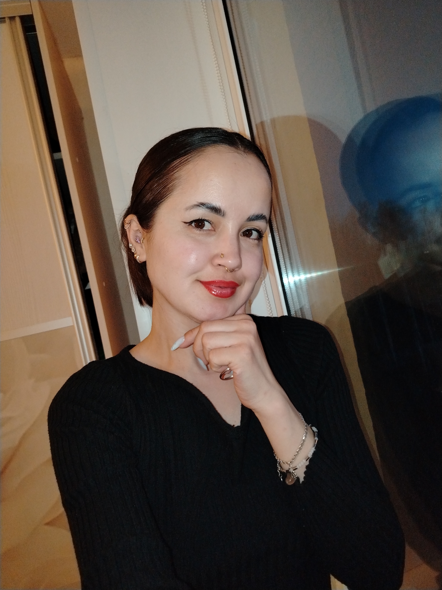 A medium shot of a woman posing indoors. She is fair-skinned with brown eyes and dark hair pulled back from her face. She is wearing a black long-sleeved V-neck shirt and a variety of jewelry: a nose ring, multiple earrings in her left ear, a bracelet on her left wrist, and a ring on her left hand. Her left hand is raised, with her chin resting on her index finger. She has makeup on, including eyeliner and red lipstick.
Behind her is a window with a light-colored frame and curtains or blinds visible at the top. A blurry reflection of a person is visible in the windowpane.
The city of Noviy, Russia, cannot be verified from the image.