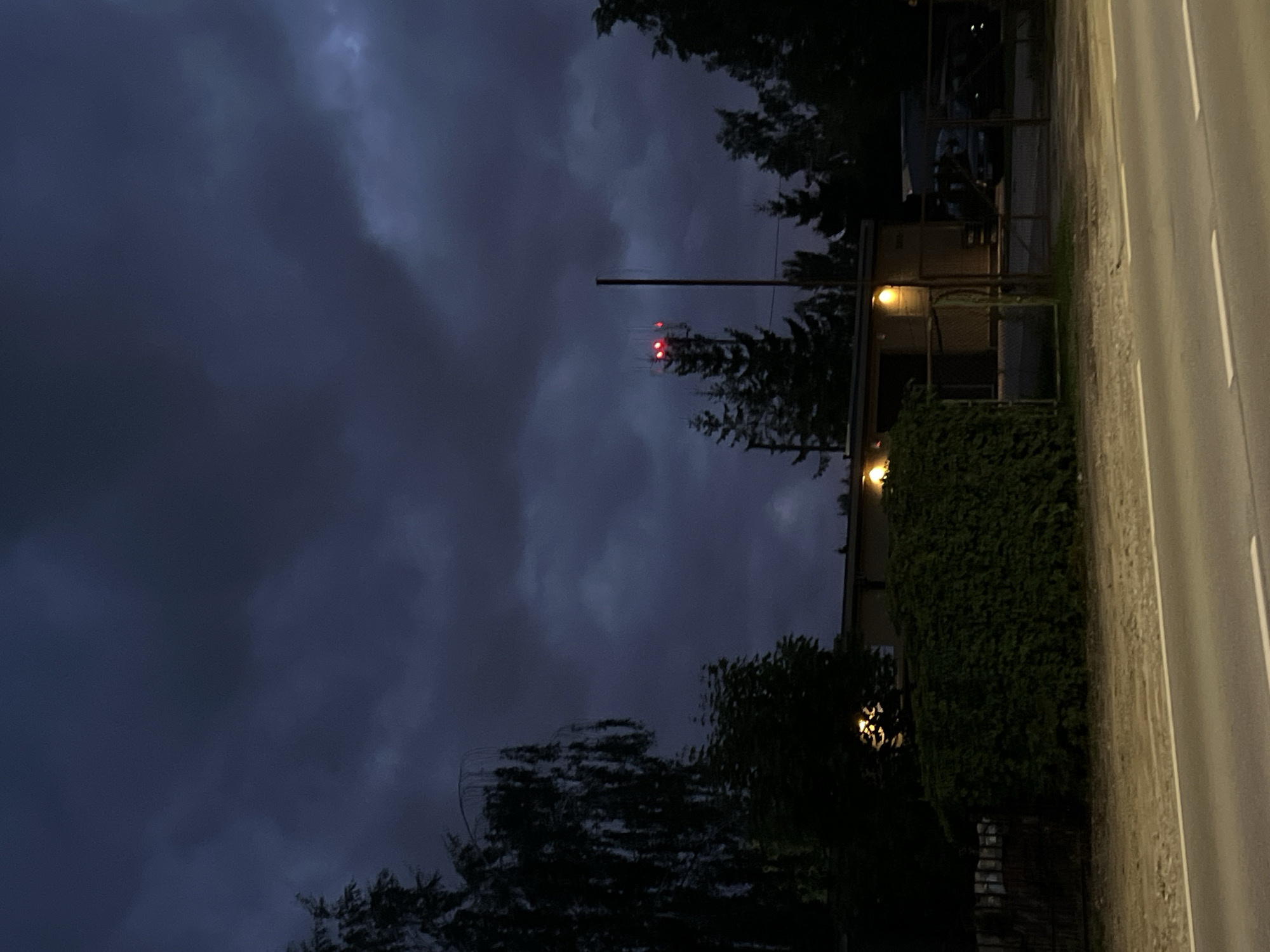 The image depicts a street scene at dusk or night in Vilnius, Lithuania. The sky is dark and cloudy, with dramatic formations of deep blue and grey. In the middle ground, a low, light-colored building with two illuminated windows is visible, partially obscured by a dense, dark green hedge. A metal fence runs along the front of the property. To the right of the building, a tall, thin pole stands, topped with what appears to be a communication antenna adorned with three red blinking lights. Behind the building and to the right, several trees, including some conifer species, are silhouetted against the dark sky. The foreground shows a paved road with white lane markings and a dirt shoulder. The overall atmosphere is dim and slightly ominous due to the dark sky and low light conditions. No people or vehicles are clearly identifiable.