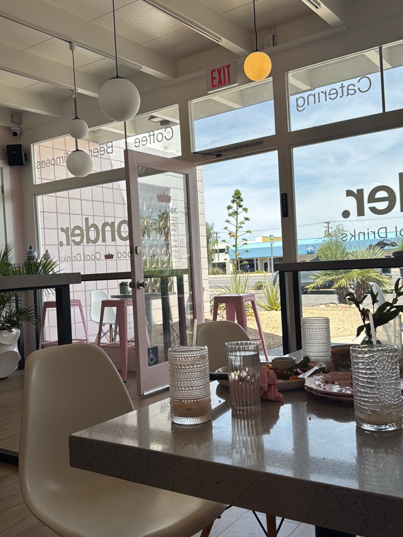 The image depicts the interior of a cafe or restaurant. A table is in the foreground, with plates and glasses on it. The table is a light gray color with a speckled pattern. A white chair is partially visible at the table. Beyond the table is an open doorway with a pink frame, with a handicap sign on the door. Outside the doorway, there are more tables and chairs. The room is brightly lit, with large windows offering views of the outside. Through the windows, trees and buildings are visible. Based on the presence of "Wonder" on the window in reverse and "Catering" on another window in reverse, the cafe is "Wonderland" located in the city of Palm Desert, United States. A sign that says "Exit" is above the door.