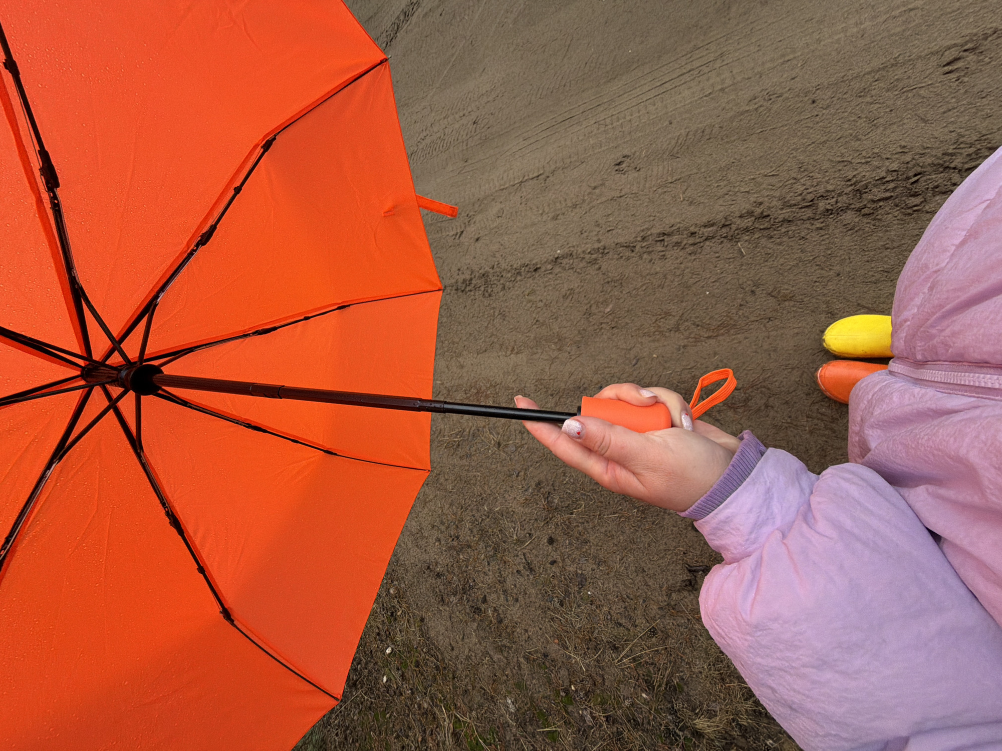A person wearing a light pink jacket and yellow and orange rubber boots is holding an orange umbrella. The ground is muddy and wet, and there are tire tracks visible. The photo was taken in Chaikovsky, Russia.