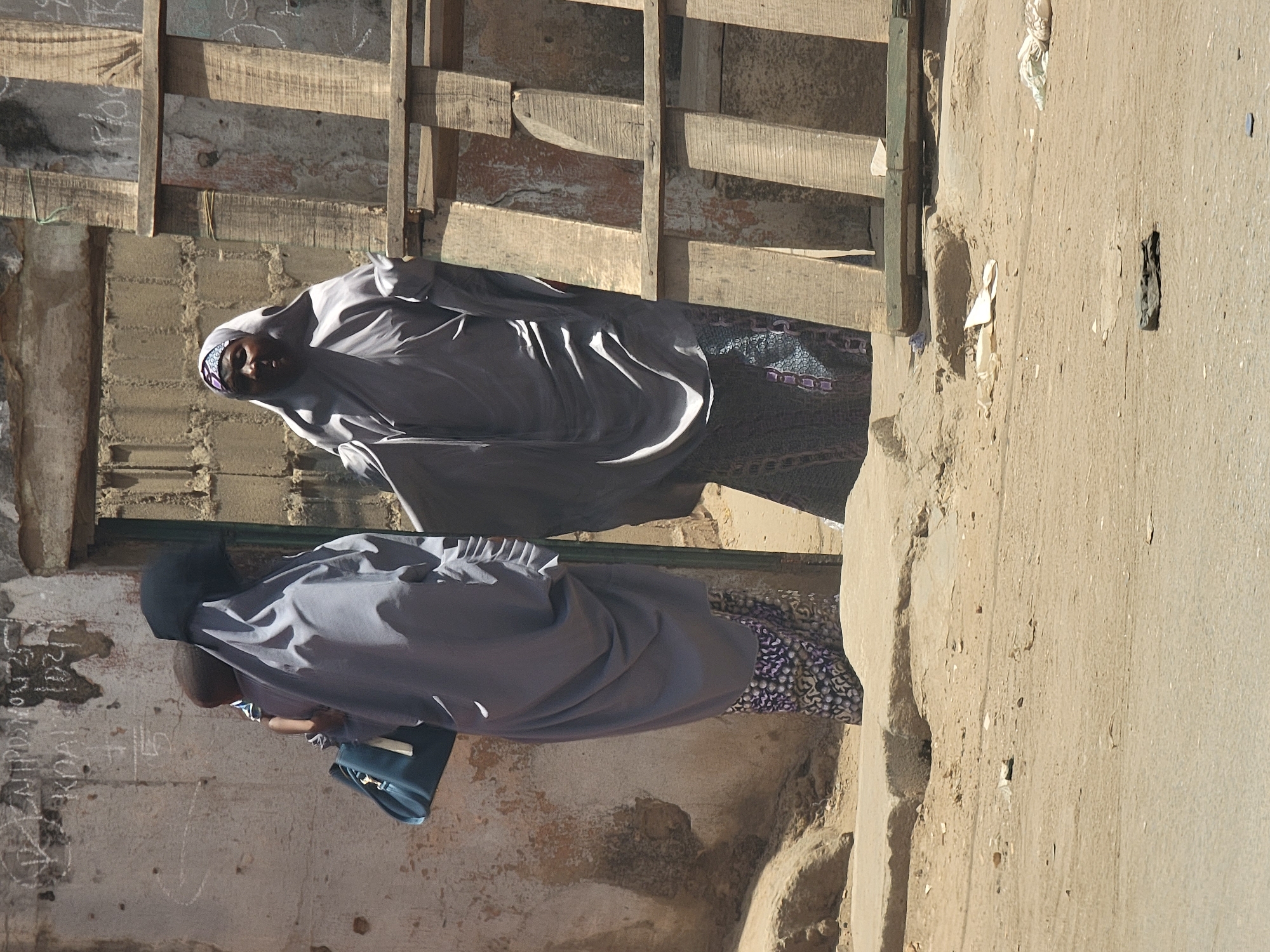 In Maiburiji, Nigeria, two women dressed in similar grey abayas and head coverings walk on a sidewalk next to a building under construction. The building has wood supports and a rough concrete facade. One woman carries a blue bag. The scene is lit by sunlight.