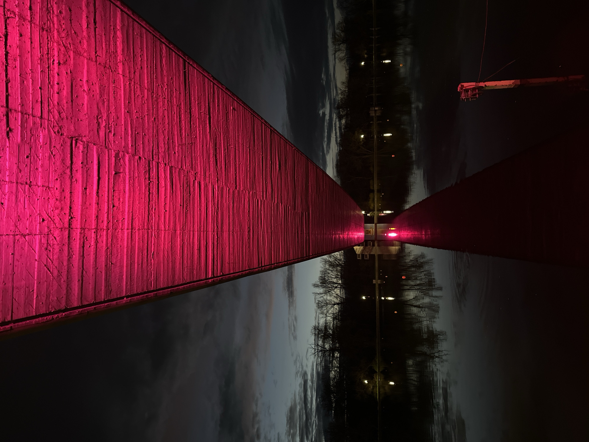 The image depicts a distorted view of a cityscape, likely taken at night near a body of water. A large, concrete structure is illuminated with bright pink light on the left side of the image. A reflection in the water is visible on the right, showing a line of lights along a road or path, as well as trees and the sky. A pole-like structure with attached hardware is visible at the top right. The horizon line is ambiguous due to the reflection and distortion, but the image gives the impression of a mirrored scene. There are no people visible. Based on the prompt, this location is in Plovdiv, Bulgaria.