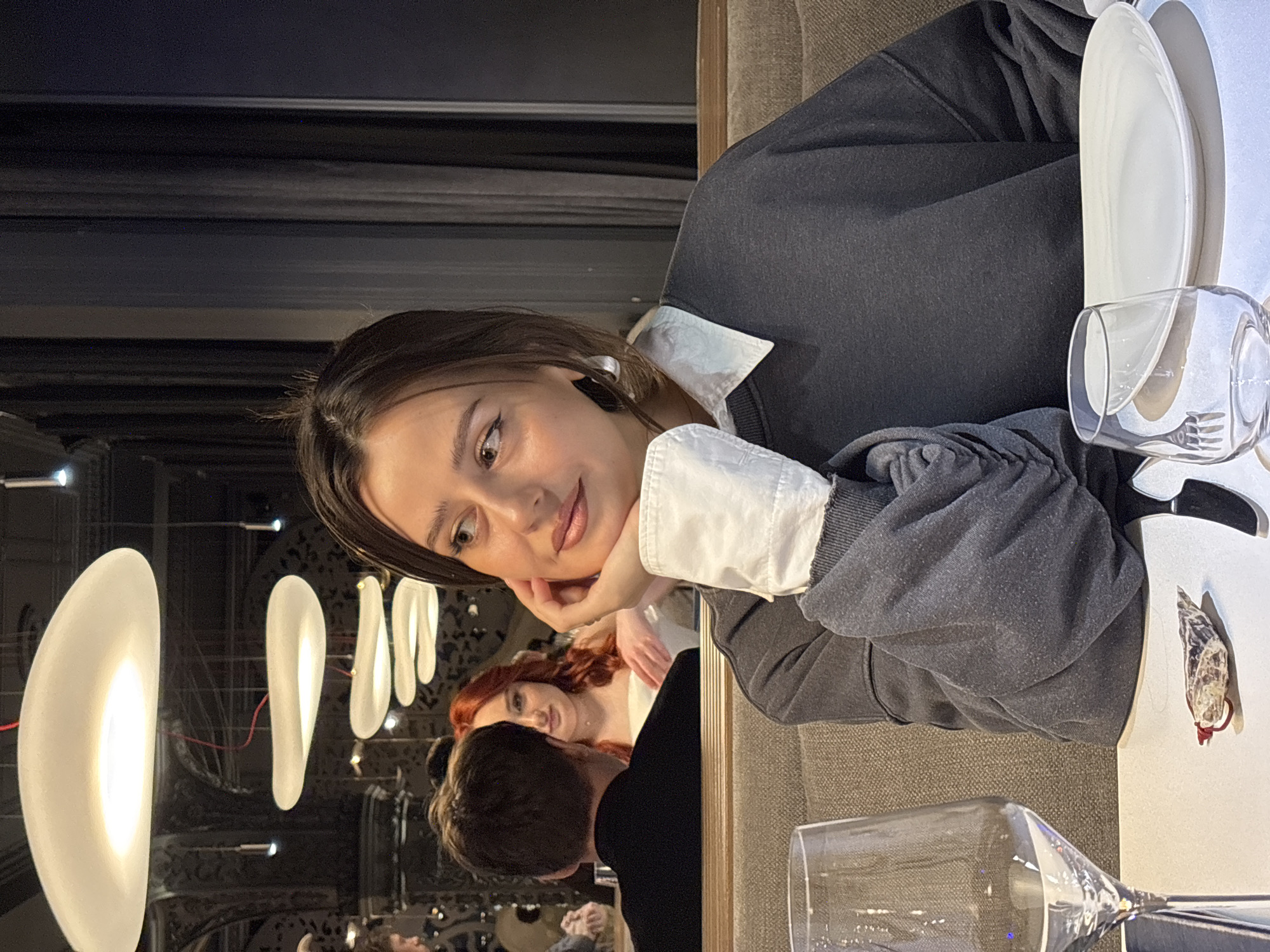 The image depicts a young woman seated at a table within Tunguska Restaurant, located at Mira Avenue, 19, Krasnoyarsk, Russia. The woman, with medium-brown hair and light skin, is positioned slightly to the left of the frame's center. She is wearing a dark grey long-sleeved sweatshirt over a white collared shirt, with the white collar and an extended cuff visible. Her left hand supports her chin, thumb along her jawline, and she looks to her right with a calm expression, light makeup including winged eyeliner visible. A silver-colored hoop earring is present on her left ear.
On the white table in front of her are two empty drinking glasses—one stemmed wine glass on the left and a stemless tumbler further right—along with a white, oval-shaped plate and a black-handled knife. A small, reddish-brown, textured object with a thin red string is positioned between the wine glass and the knife. The seating appears to be a light beige upholstered booth.
In the background, other patrons are visible. To the main woman's left, partially obscured, is the back of a man's head wearing a dark shirt. Further back, a woman with long, wavy reddish-orange hair is seen looking towards the foreground, wearing a white strapless garment, suggesting a formal or semi-formal event.
The restaurant's interior is dimly lit but features distinctive modern decor. Several white, organic-shaped pendant light fixtures hang from the ceiling, some illuminated, casting a soft glow. Dark curtains are visible on the right side of the frame, and some arched architectural details are present in the deeper background, indicating a refined dining environment.