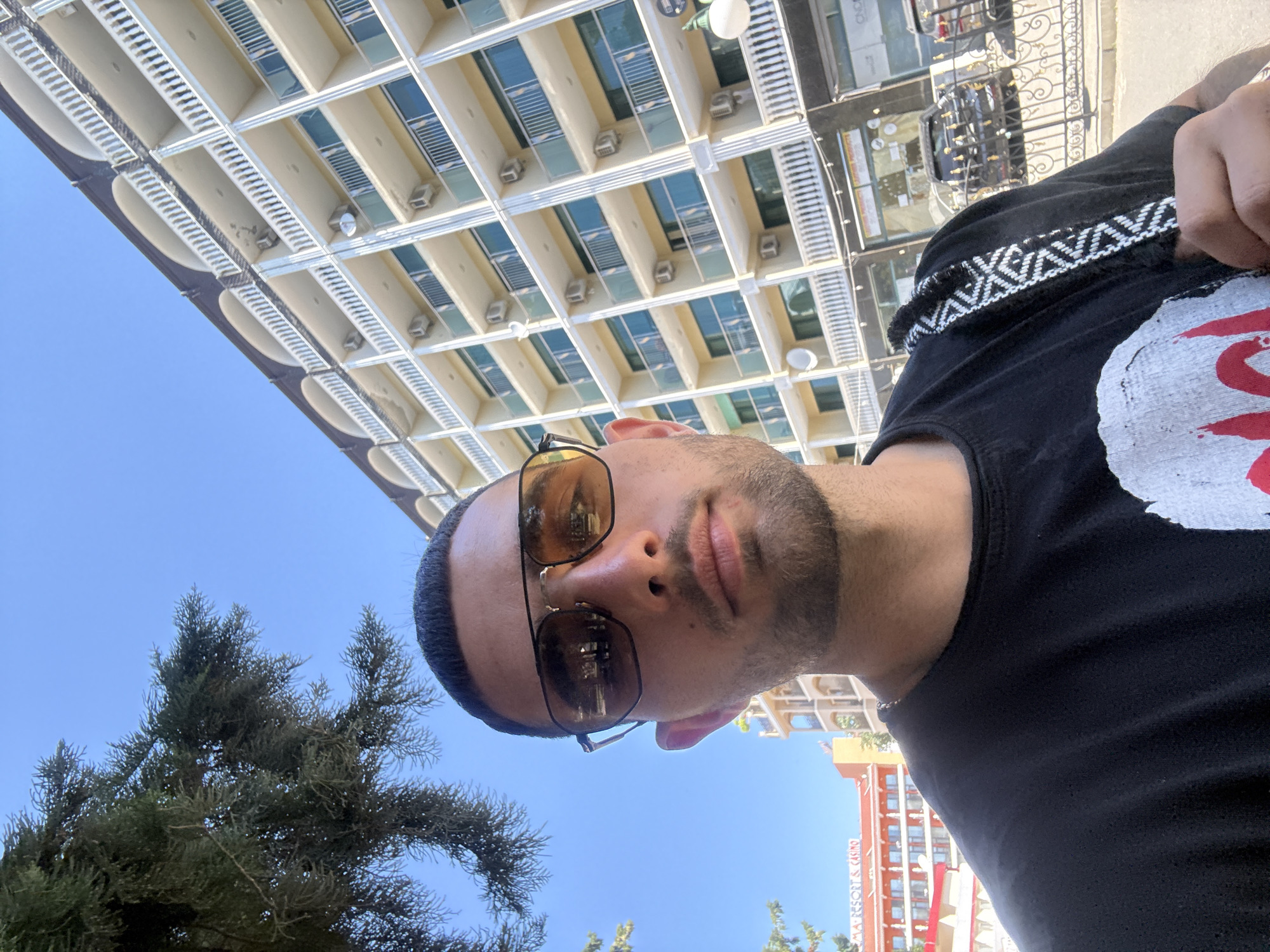 A man wearing sunglasses and a black t-shirt is taking a selfie outdoors in front of a hotel building. The man has short dark hair and stubble. He is looking directly at the camera. Behind him, a large, light-colored hotel building with balconies and air conditioning units is visible against a clear blue sky. A tree with green foliage is to the left of the man, partially obscuring the sky. In the background, other buildings and a street with parked cars can be seen. The man has a patterned strap over his shoulder, which appears to be part of a bag. This scene is located in the city of Nesebar, Bulgaria, a popular resort town on the Black Sea coast.