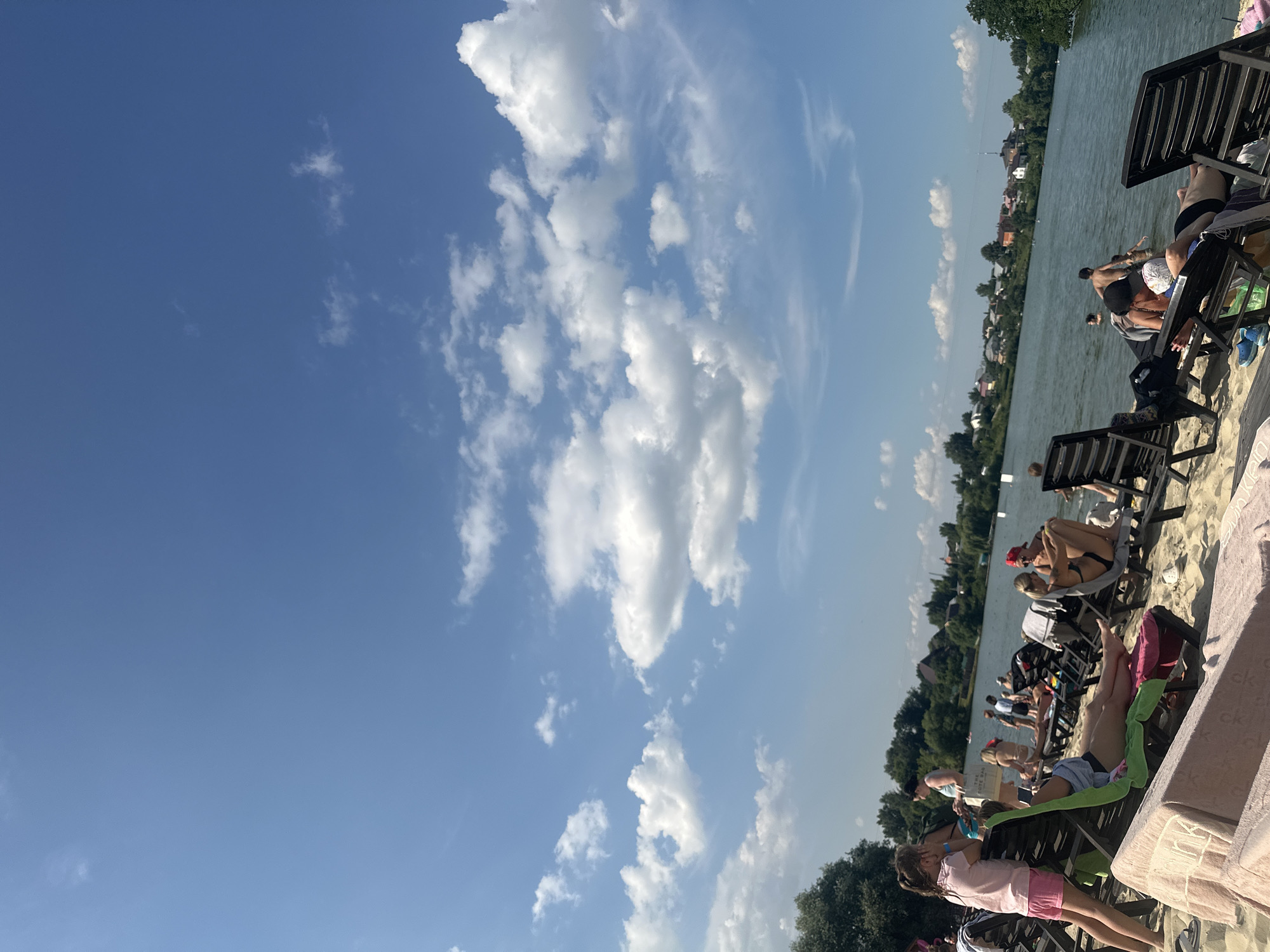 This media file captures a sunny day at a beach resort on a lake, likely in Dal'nie Prudishchi, Russia. The image is oriented vertically, with the sky dominating the left half and the beach and lake filling the right. Fluffy white clouds dot the clear blue sky, suggesting pleasant weather, likely midday due to the bright sunlight casting shadows and illuminating the scene.
The foreground features a sandy beach with several lounge chairs occupied by people. In the middle ground, the calm lake is visible, with a line of trees and buildings along the opposite shore, indicating a populated area or small town. Several individuals are seen relaxing in the chairs, some in swimwear, enjoying the warm weather. A few people are also visible in the water, perhaps swimming or wading. The overall mood seems relaxed and leisurely, typical of a summer day at a recreational spot.
One lounge chair in the foreground has a pink blanket with the word "ck" repeated on it. The scene depicts a typical summer day with people enjoying outdoor leisure activities by a body of water.