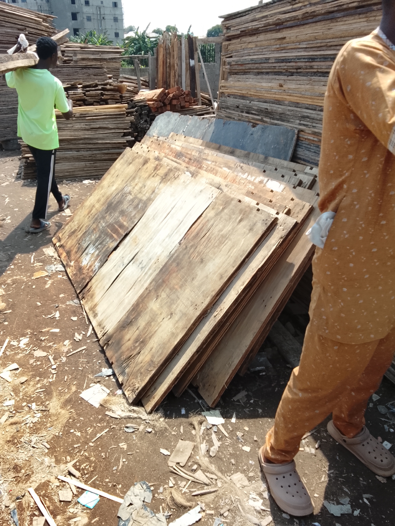 In the city of Abuja, Nigeria, a young Black male, wearing a bright green t-shirt and black pants, carries a large piece of wood over his right shoulder. He walks across a dusty, debris-strewn lot filled with stacks of lumber of various sizes, appearing to be a lumberyard or construction material storage area. To the right, another person, partially visible from the waist down, wears an orange patterned garment and brown Croc-style shoes, standing near a large stack of wooden planks. The background shows more stacks of lumber, some metal structures, and a multi-story building under construction, with some vegetation visible. The lighting suggests it is daytime, with shadows indicating a sunny environment. The ground is uneven and covered with wood scraps, dust, and some scattered plastic debris.