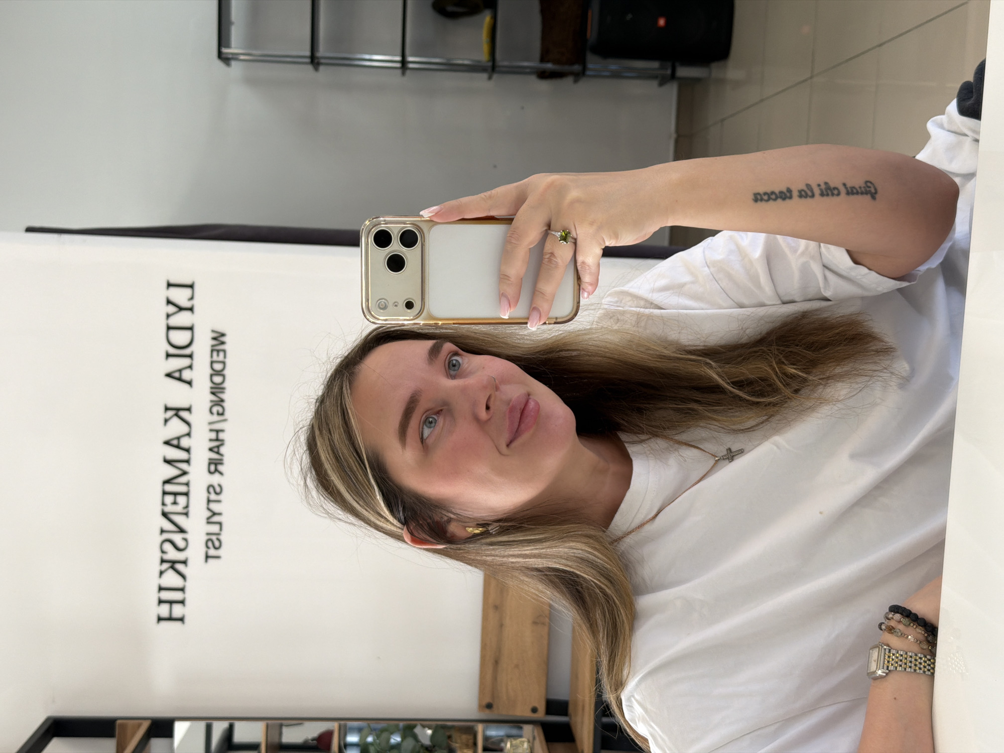 The media file captures a mirror selfie taken by a woman in what appears to be a salon or studio, located in Chaikovsky, Russia. The woman, with light eyes and long, highlighted hair, is smiling subtly as she holds an iPhone in a gold-rimmed clear case to take the picture. She is wearing a white t-shirt, a small cross pendant necklace, and has a nose piercing.
Notable details include a prominent ring with a green gemstone on her left ring finger, and on her right wrist, a watch with a gold and silver band worn alongside multiple beaded bracelets. Her fingernails are manicured with French tips. A tattoo is visible on her right upper arm, reading "quasi ita al toccata" (Italian for "almost touched by it") when un-mirrored.
The background consists of a white wall featuring a mirrored sign that, when deciphered, likely reads "HIKMEHKIK STYLISTS | HAIR | WEDDING MEDIA," indicating the establishment is a hair or wedding services salon. Parts of shelving units are visible at the top and bottom edges of the frame, reinforcing the indoor commercial setting. The lighting is bright and even.