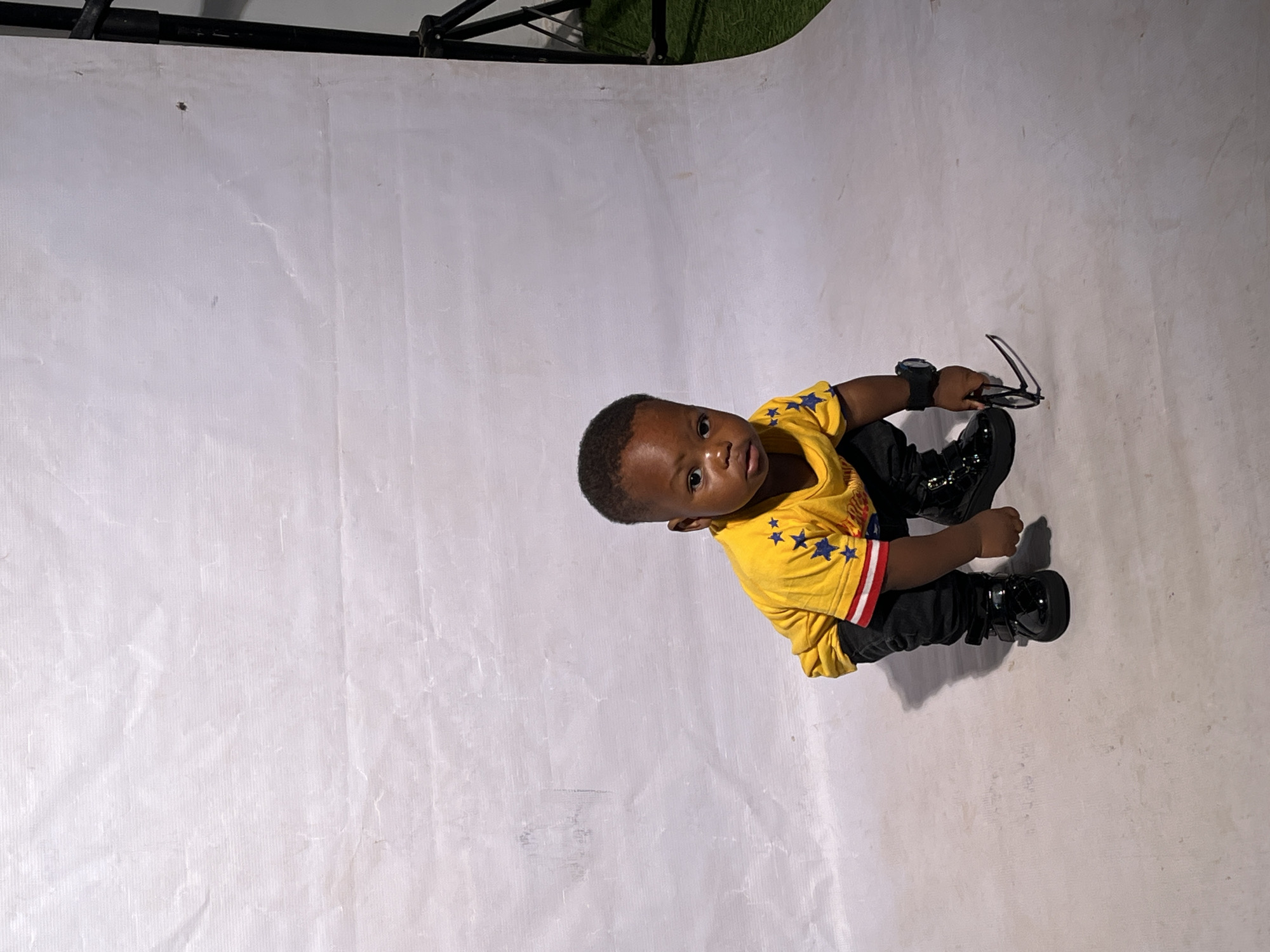 A young child is captured in a low-angle, full-body shot, crouching on a light-colored, textured floor. The child, appearing to be of African descent, is wearing a bright yellow t-shirt adorned with blue stars and red and white striped sleeves. Dark pants and shiny black boots complete the outfit. The child is holding a pair of dark-rimmed glasses in their right hand, which is near the floor. A dark wristwatch is visible on the child's left wrist. The background consists of a large, crumpled white fabric sheet, suggestive of a studio setting. To the right of the frame, a black tripod stand is partially visible, along with a hint of green turf, possibly indicating an outdoor or mixed-environment setup. The lighting appears to be artificial, creating soft shadows. The child's gaze is directed towards the viewer with a neutral expression. The scene evokes a sense of posed portraiture. The context of Barakin Akawo, Nigeria, is not visually discernible from the provided image.