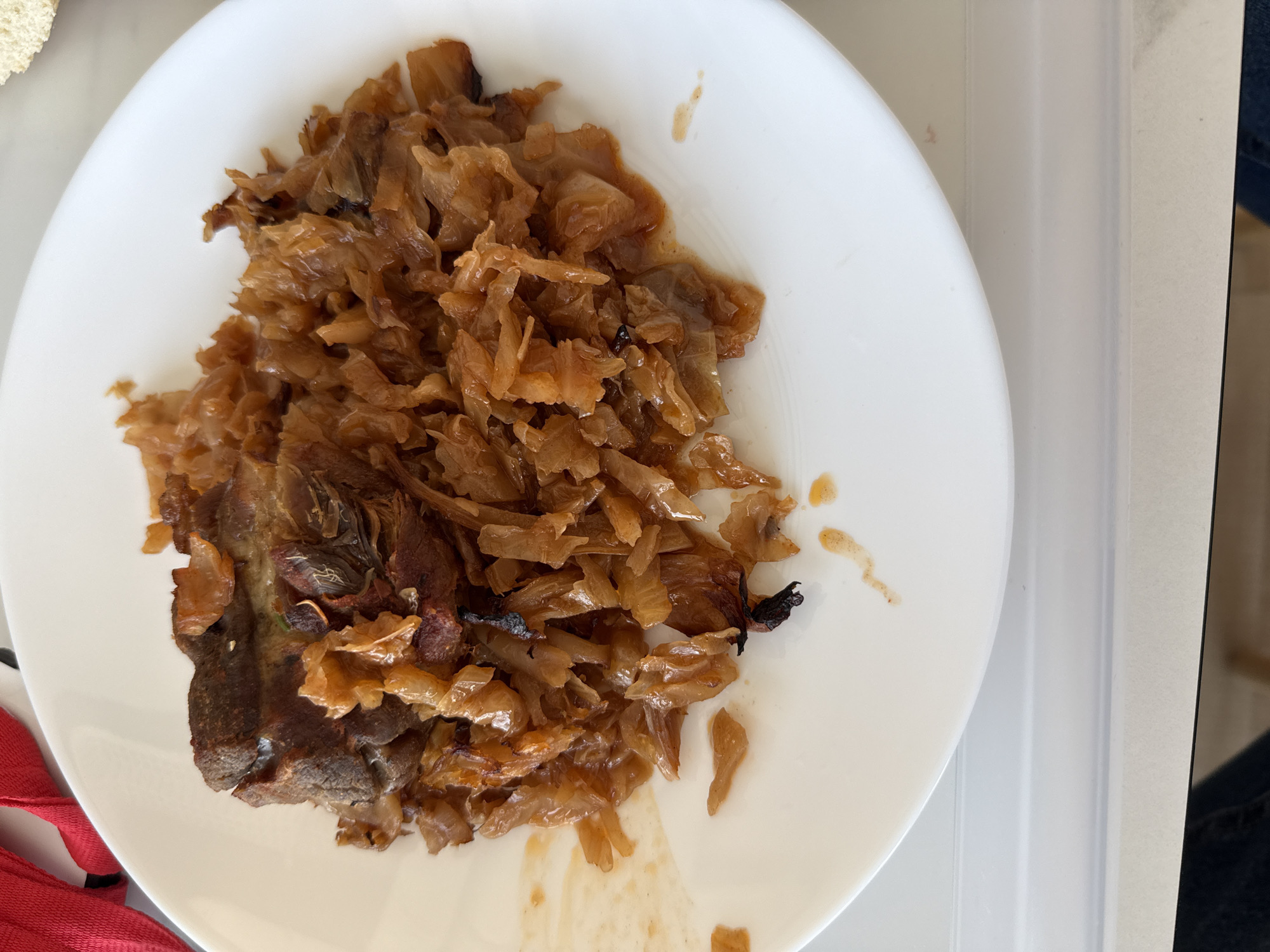 A top-down shot depicts a white plate holding cooked food. The food consists of a portion of dark meat on the left side of the plate, which appears charred in some areas. Next to it, and partially covering the meat, is a heap of sauteed or stewed cabbage with a light brown coloration. There are some stray pieces of cabbage and some oily residue spread on the plate. The plate is situated on a surface with visible segments of what may be a white countertop, and a hint of a red cloth is visible on the bottom left. To the right is clothing or furniture in neutral tones. The scene does not contain any visual cues that indicate it is located in the city of Sofia, Bulgaria.