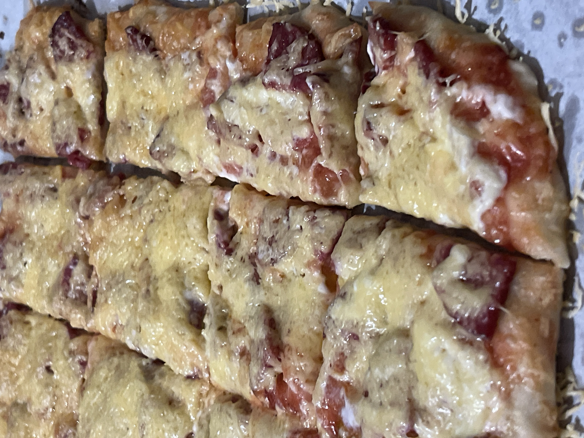 A close-up, overhead shot shows a rectangular pizza that has been cut into multiple square slices and arranged in a grid pattern. The pizza has a golden-brown crust, topped with melted cheese, tomato sauce, and slices of pepperoni. The lighting is even, highlighting the textures of the melted cheese and toppings. The background is a muted gray surface.