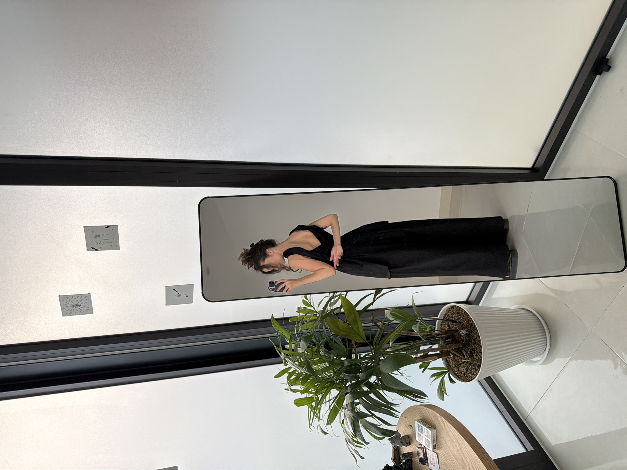 The image captures an indoor scene, brightly lit, likely during the daytime. The primary subject is a woman, reflected in a full-length, black-framed mirror, taking a mirror selfie. She has dark hair styled in a messy bun or updo and is wearing a elegant black, backless dress with a full skirt. Her body is slightly turned, and she is holding a dark-colored smartphone, possibly an iPhone, in her left hand, looking at its screen.
The setting appears to be a modern, minimalist space with large windows or glass panels that feature black frames along the top and left side (when viewing the image in its correct, upright orientation). To the left of the mirror, on a light-colored wall, are several small square stickers or papers with faint, abstract graphics. The background reflected in the mirror is a plain, light-colored wall.
In the foreground, a large potted plant with lush green leaves rests in a white, ridged pot with a matching saucer on a light-colored, glossy tiled floor. Part of a round wooden table, laden with small, indistinct objects like books or decorative items, is visible next to the plant. The floor shows reflections of the light. On the woman's phone screen, reflected in the mirror, the number "5.0" is visible, possibly indicating a camera setting or timer. The overall atmosphere suggests a contemporary space, consistent with locations found in Đà Nẵng, Vietnam.