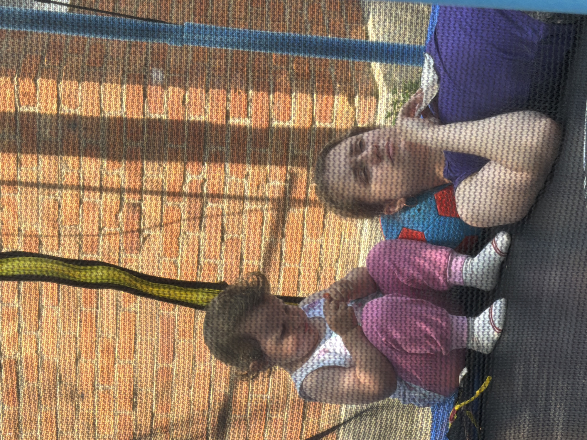 In Komsomolskiy, Russia, two individuals are captured in an outdoor setting, possibly a backyard, during daylight. A young child, seated, is wearing a sleeveless white top with a floral pattern and pink pants with white socks. The child is focused on something in their hands. Beside the child, an adult reclines, with their head resting on their hand. The adult is wearing a purple top. They are positioned on a dark-colored surface, likely a trampoline mat, which is part of a larger trampoline structure with a blue frame. The background consists of a textured, orange brick wall and a green and black striped element, possibly part of the trampoline's safety netting. Sunlight casts shadows across the scene, creating a dappled effect.