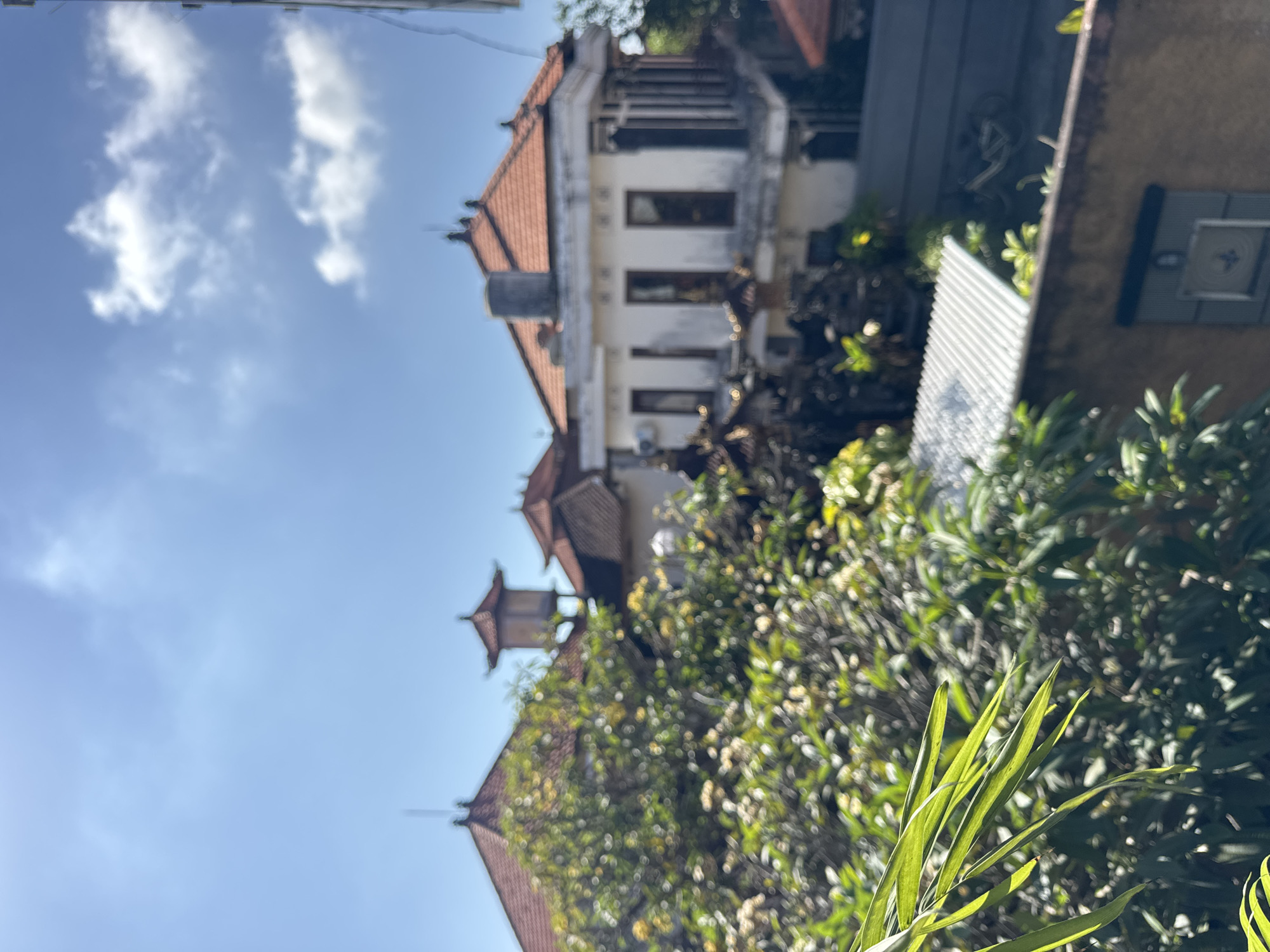 The image depicts a residential scene in Kuta, Indonesia. A multi-story building with a traditional Balinese architectural style is visible, featuring a red tile roof and white walls with several windows. Dense tropical vegetation, including trees and bushes, partially obscures the building. The sky is blue with scattered clouds. The perspective is from a low angle, looking upward. Foreground elements consist of plant leaves. The scene appears to be outdoors during daylight hours.