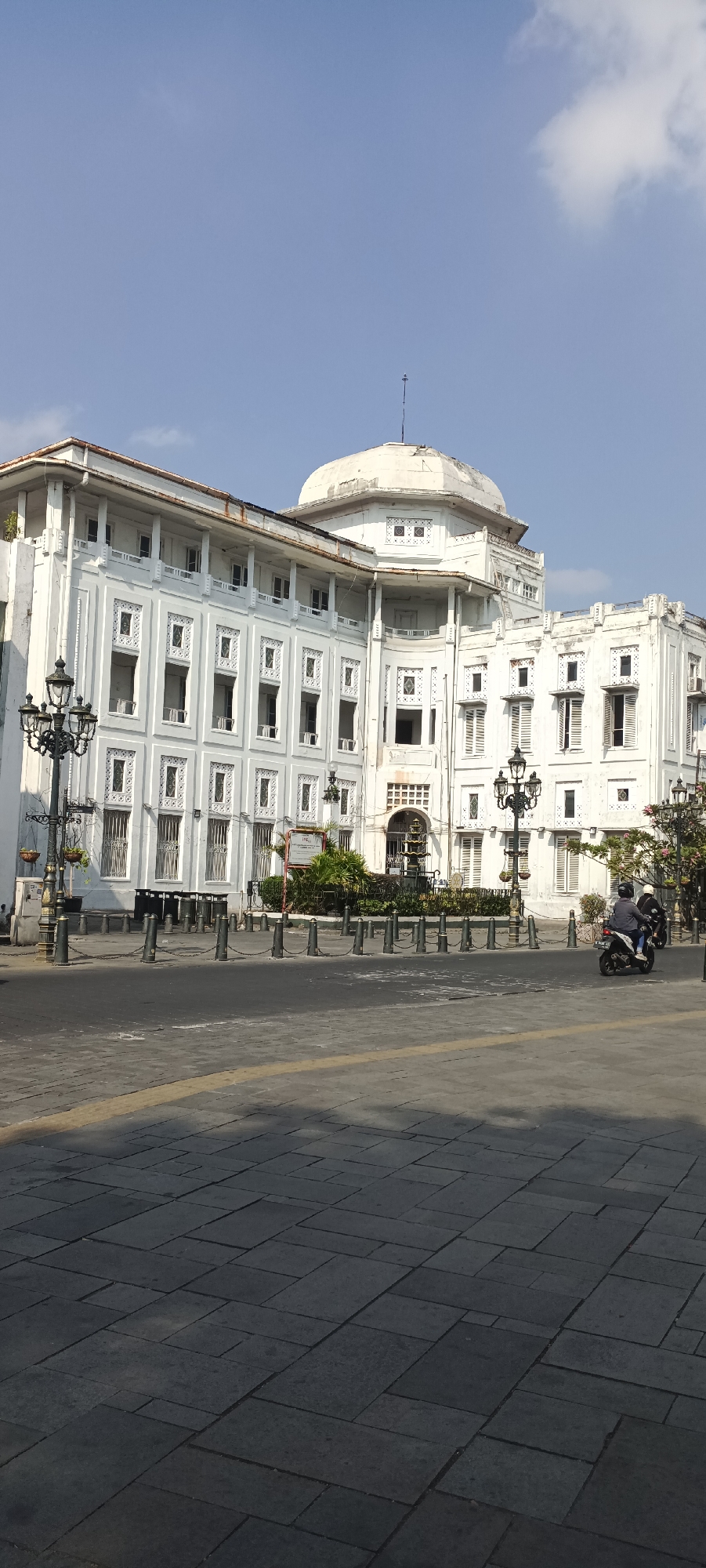 A white, multi-story colonial-era building with a domed structure on top is visible under a clear blue sky in Semarang, Indonesia. The building features numerous windows with ornate decorative panels. A paved area with patterned grey stone slabs is in the foreground, with a yellow line marking a boundary. Several black bollards connected by chains are arranged along the edge of the paved area. Two ornate black lampposts stand on the left side of the image. A motorcycle with two occupants is seen in the middle ground, moving away from the camera. Lush green plants are visible in front of the building.