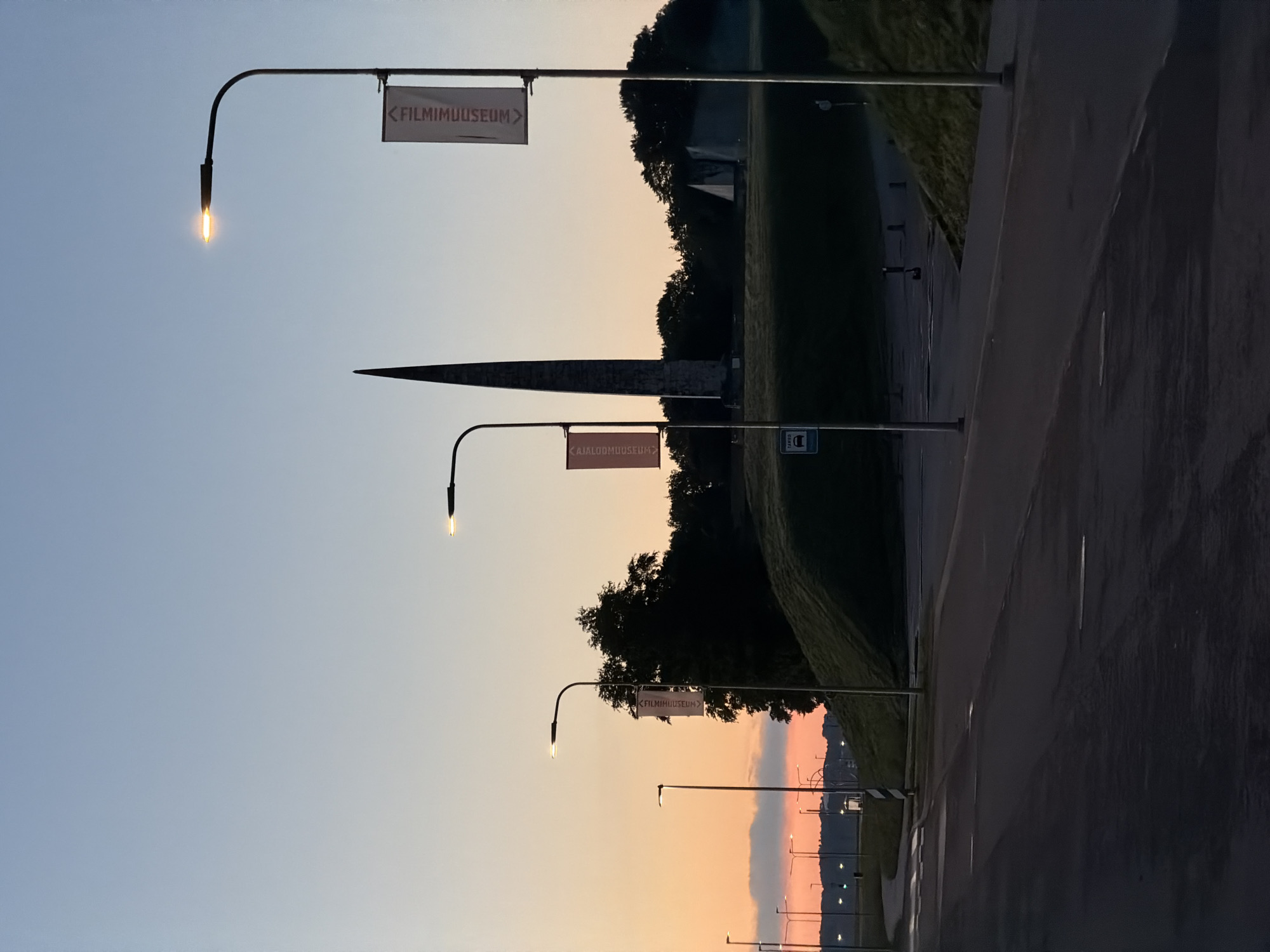 The image depicts the Pirita Marina complex in Tallinn, Estonia, at dusk. A tall, obelisk-like monument dominates the background, situated on a grassy hill. The sky transitions from a pale blue at the top to a warm orange and pink hue near the horizon, indicating sunset or sunrise. Several streetlights line a paved road in the foreground, some of which are illuminated. Two banners, one on the left and one on the right, hang from lampposts, each displaying the text "<FILMIMUUSEUM>" in white against a red background. The road appears wet, possibly from recent rain, with reflections visible on its surface. A bus stop sign is visible on a lamppost to the left of the monument. The overall scene is quiet and serene, with no people or vehicles immediately apparent. The architecture of the monument and the surrounding landscape are characteristic of this area of Tallinn.
