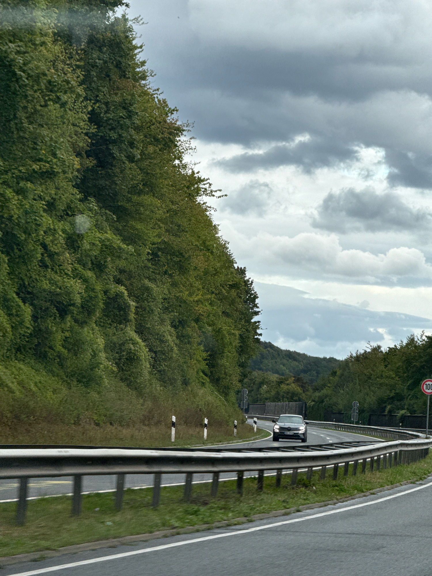 The image shows a view from a vehicle traveling on a highway in Herzberg, Germany. A silver sedan is visible ahead, driving on the same road. The highway is flanked by dense green trees and foliage on the left and a gently curving barrier on the right. The sky is overcast with dramatic gray clouds. A speed limit sign indicating 100 km/h is visible on the right side of the road. The road surface is asphalt with white lane markings.
