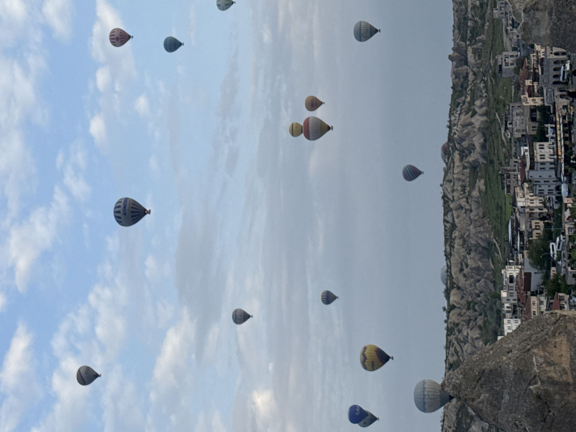 The image depicts numerous hot air balloons floating over Göreme, Turkey. In the left portion of the image, a blue sky is interspersed with white clouds. The right portion of the image shows a landscape of unusual rock formations and buildings. The buildings are small and densely packed together. A large rock formation is visible in the bottom right corner of the image.
