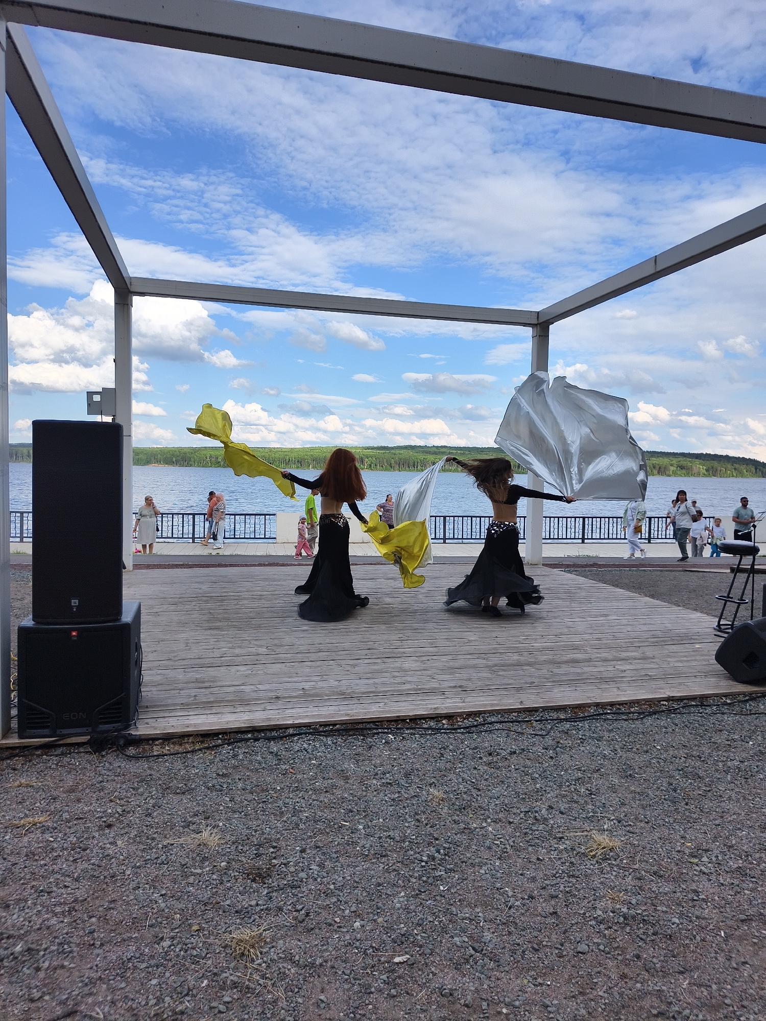 An outdoor stage with a wooden floor is set up near a body of water. Two female dancers in black costumes are performing, one with a yellow cloth and the other with a silver cloth. They are in the middle of the stage, with their backs to the camera. A large black speaker is positioned to the left of the stage. In the background, people are standing on a boardwalk, and a tree line borders the water. The sky is blue with scattered white clouds.